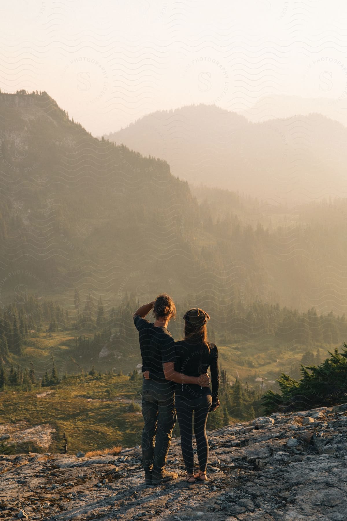 A couple standing at a cliff edge overlooking a forest with trees and mountains below