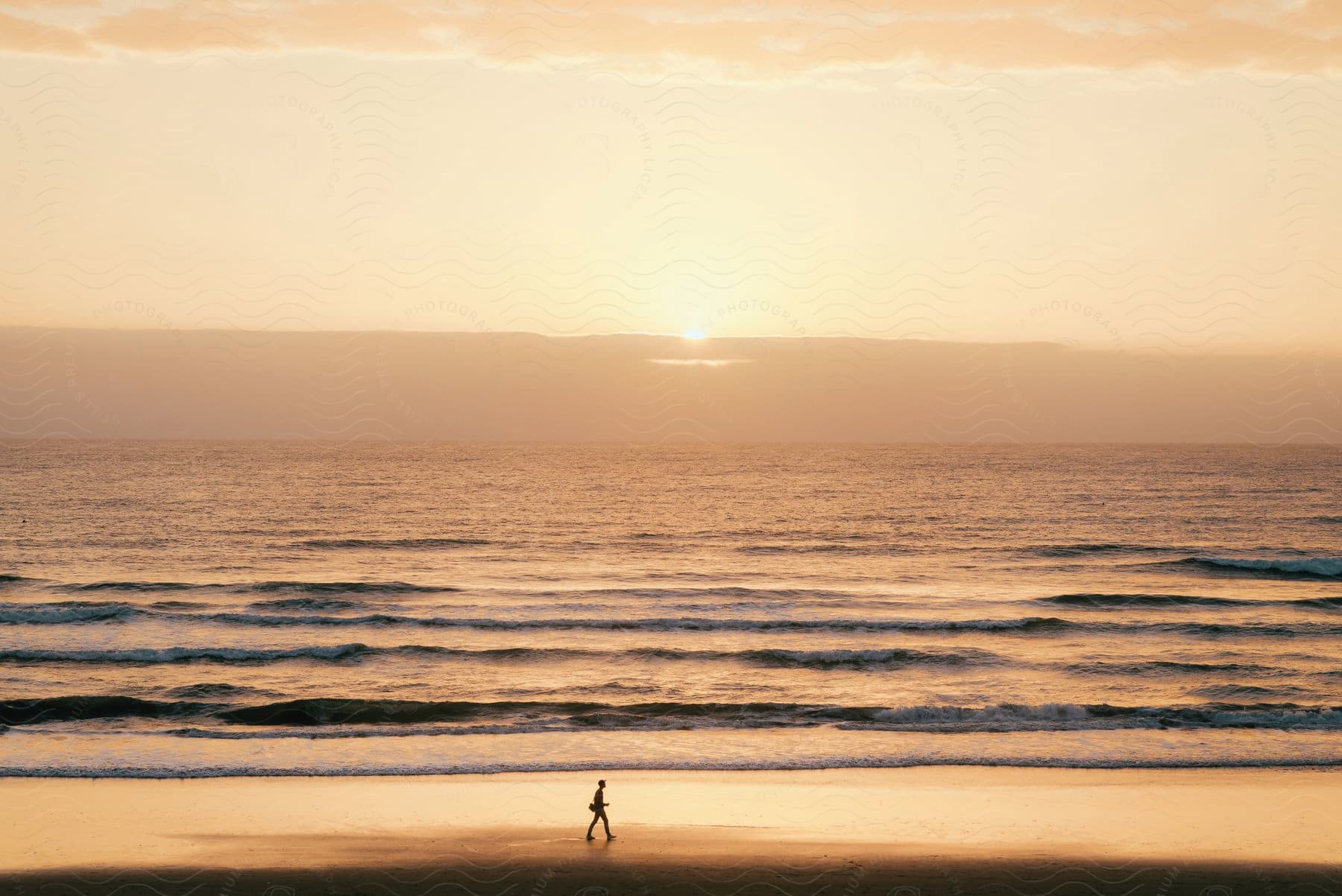 A man walks on the beach as waves roll in and the sun shines on the horizon