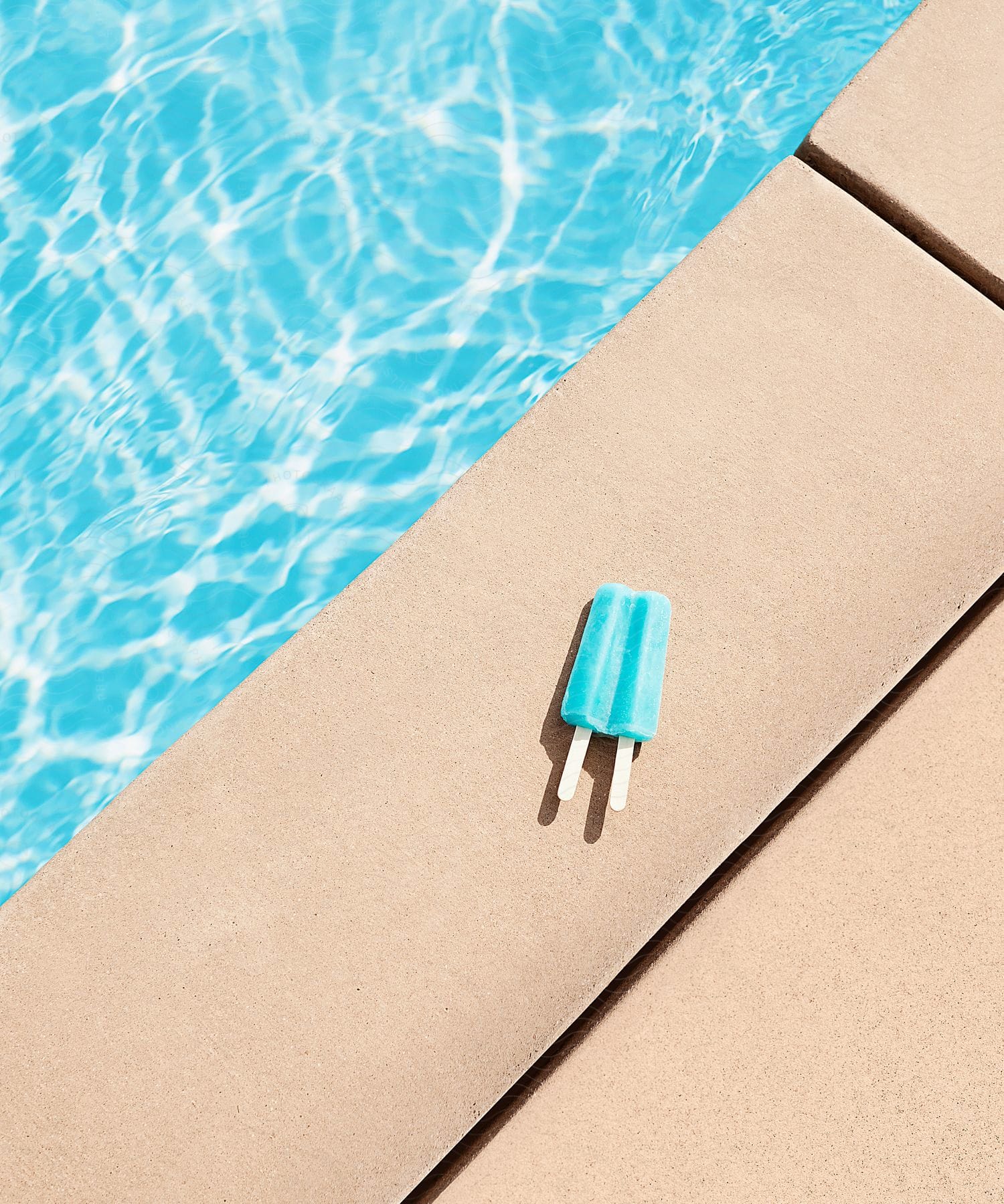 A blue popsicle is placed on the edge of a pool in the sun