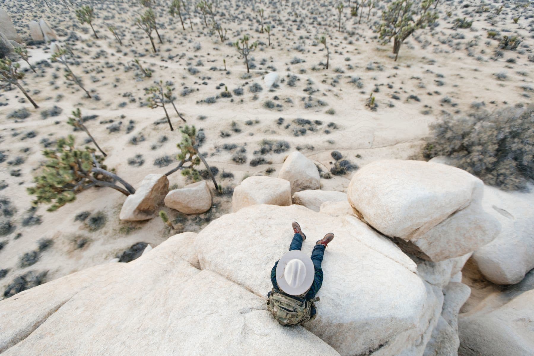 A man in his 20s30s wearing a cowboy hat backpack jacket and jeans sits on a rock in the desert observing his surroundings