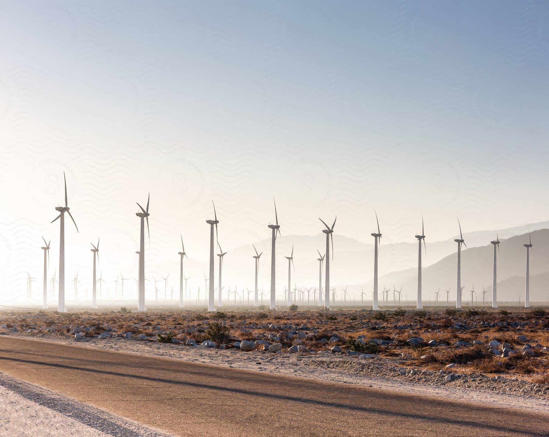 Many wind turbines stretch across the roadside with mountains in the distance under a hazy foggy sky