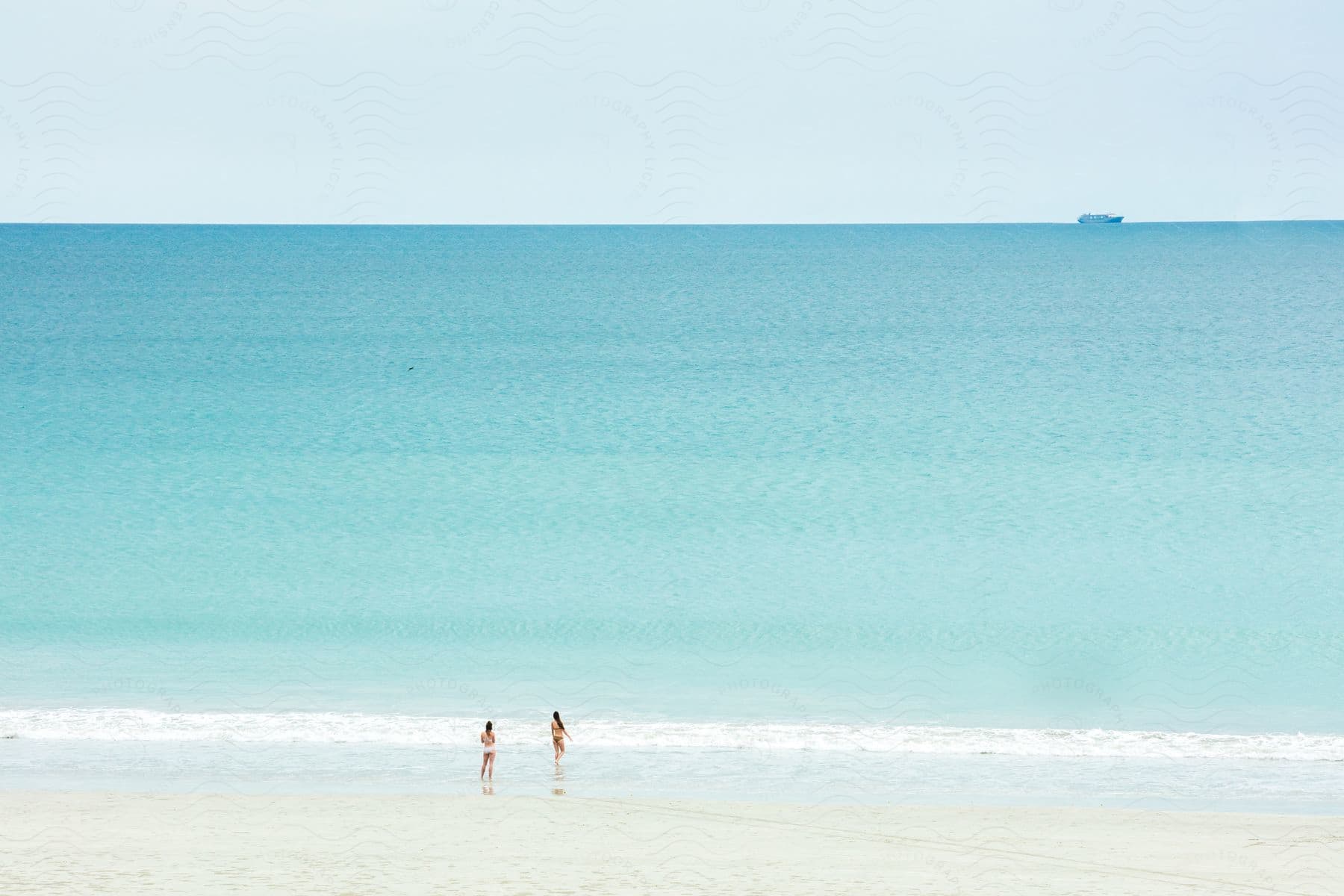 Two women walk into the water on a beach as waves roll in and a ship sails on the horizon