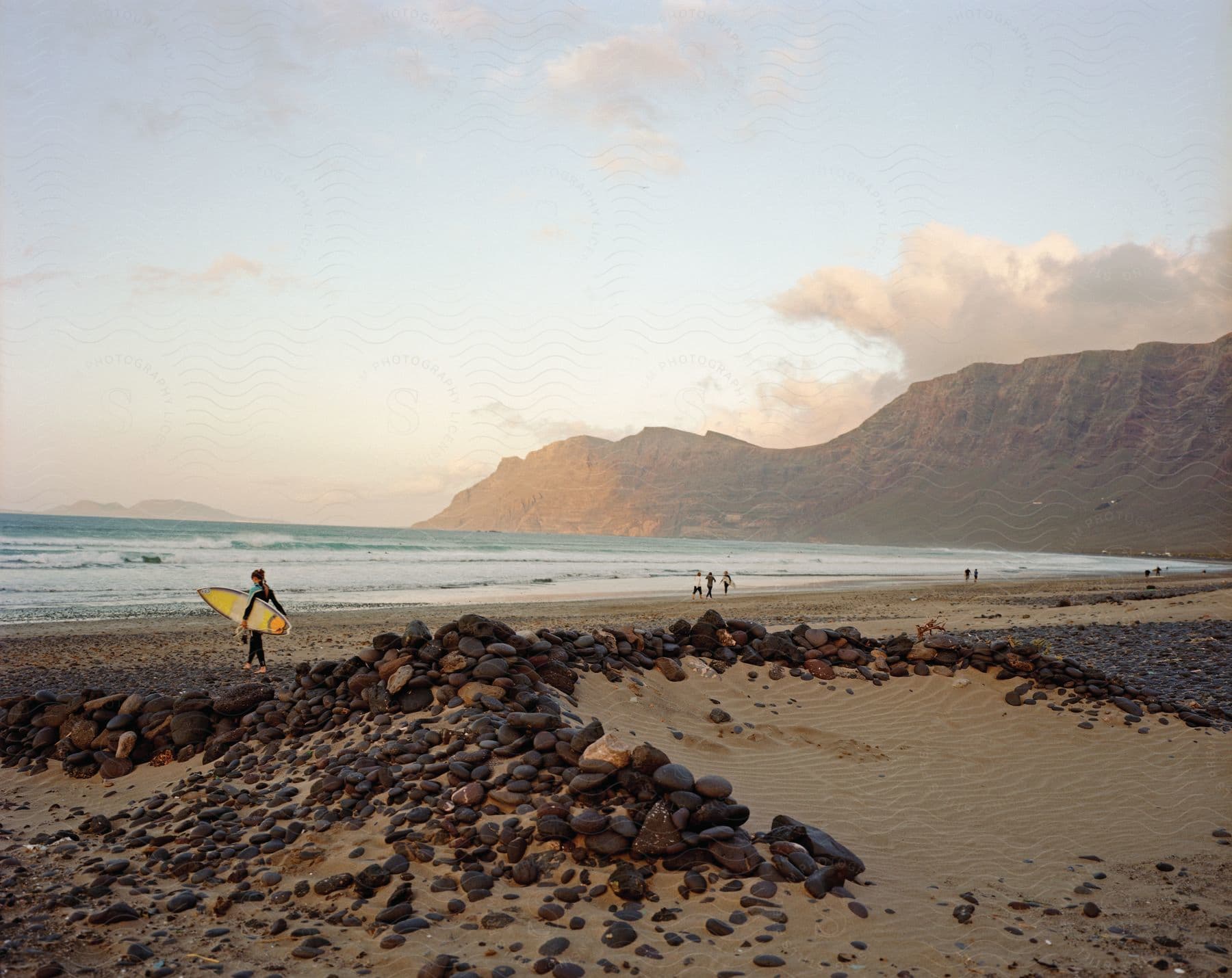 A beach at dusk with rocks on the shore a mountain in the background and groups of people walking on the beach one holding a surfboard