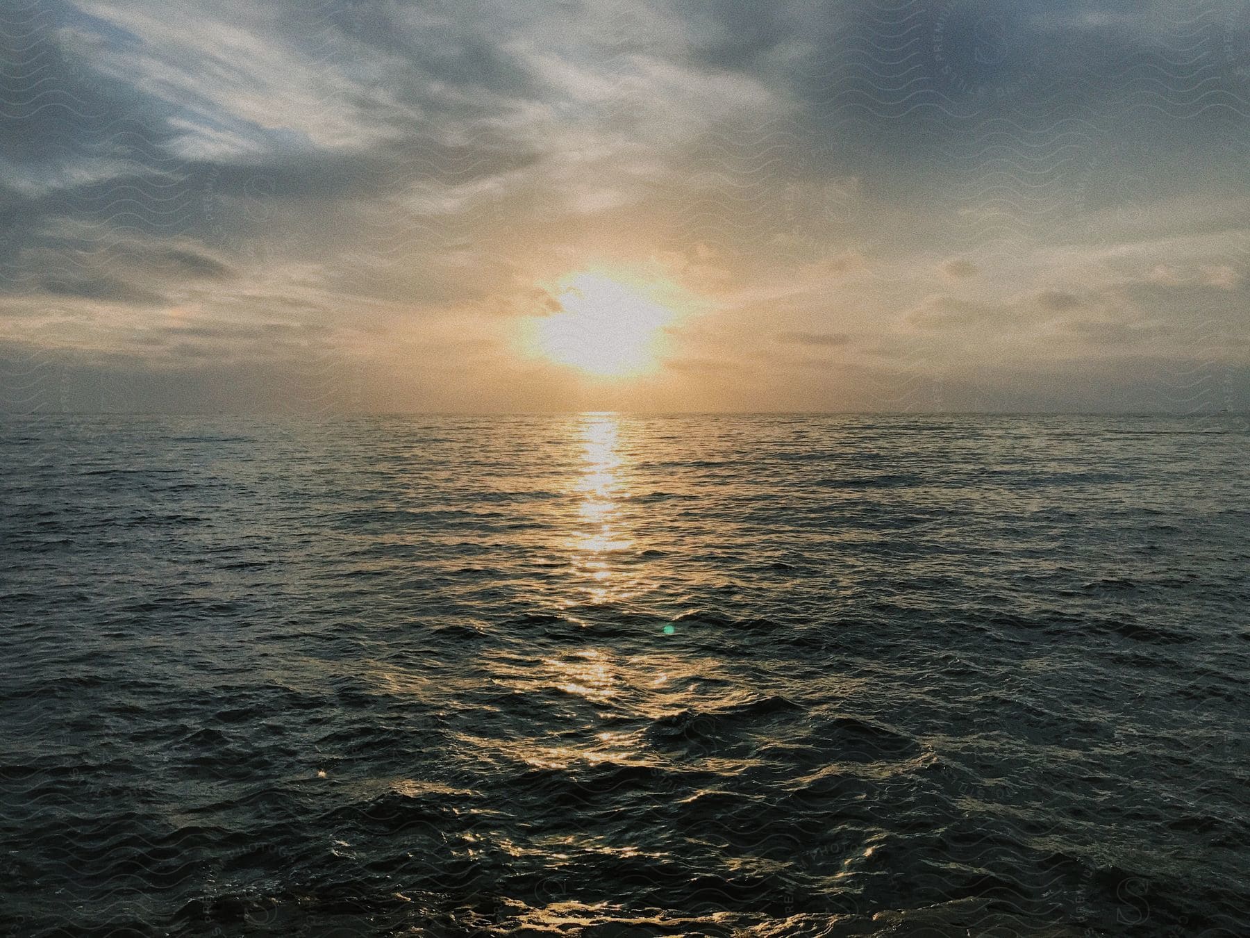 The sun sets in a cloudy sky as light reflects on the ripples and waves in the water