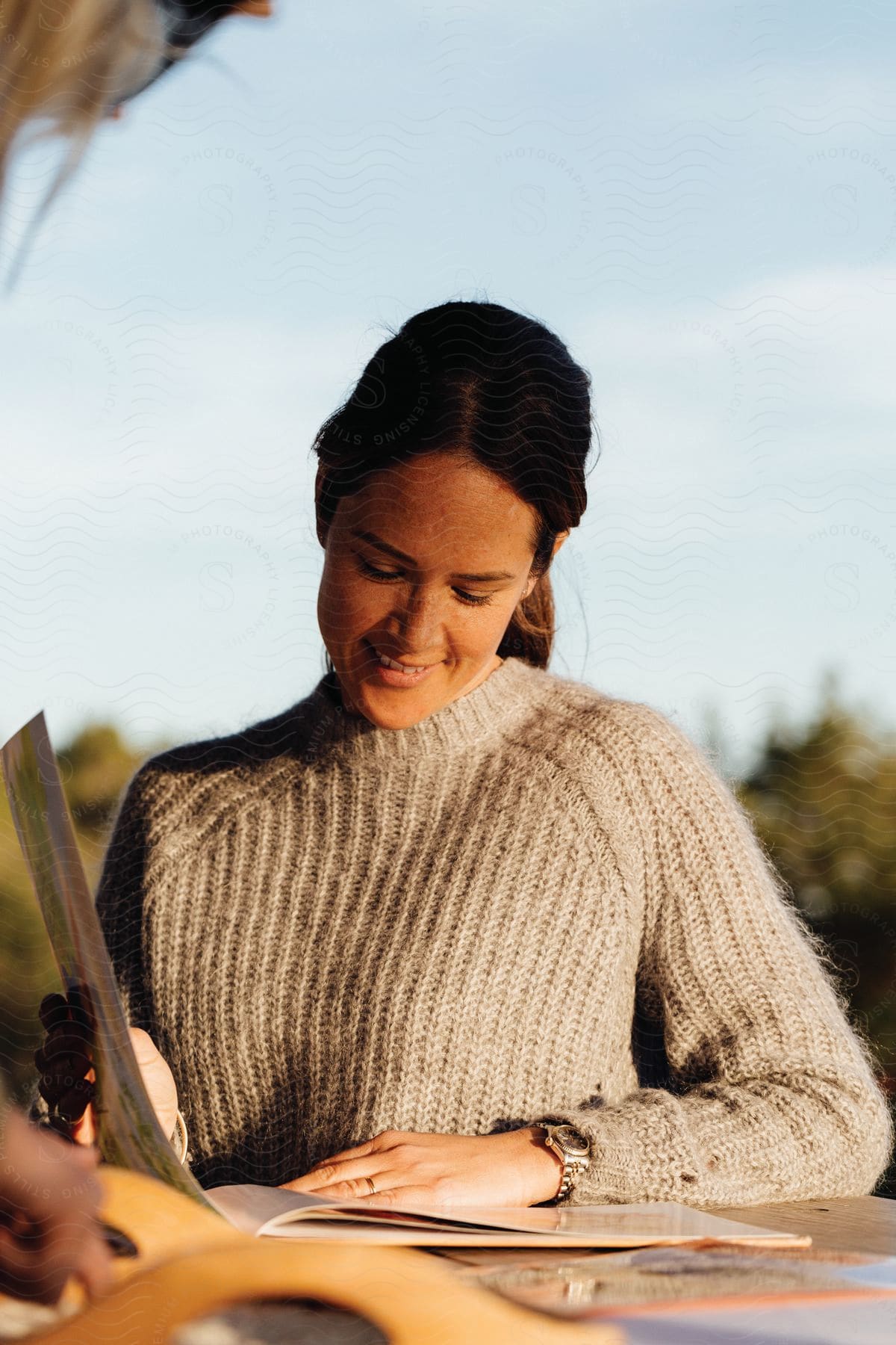 A woman wearing a tan sweater looks down at a book outdoors