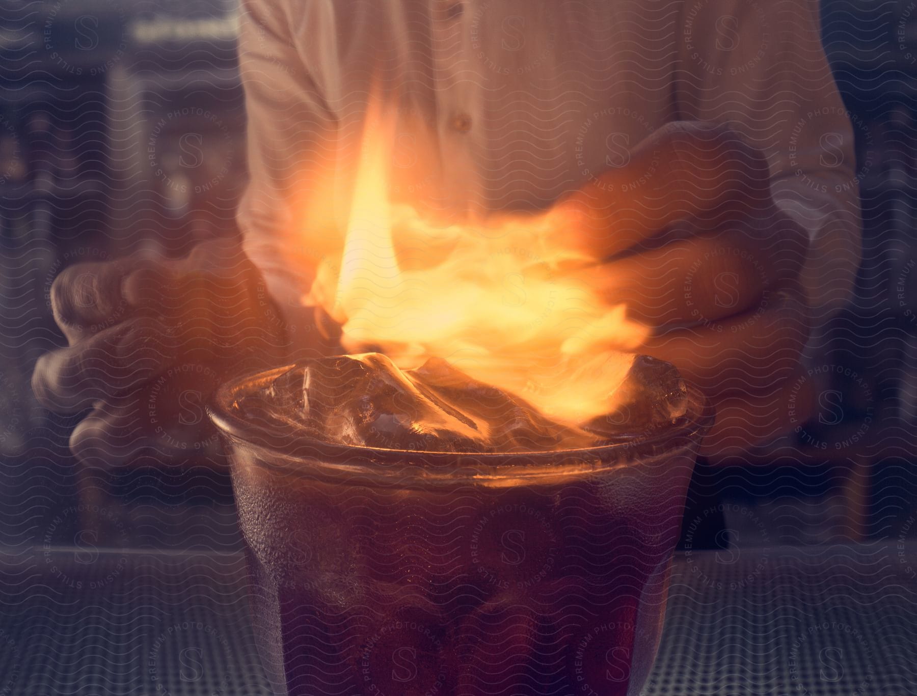 A cooks hands setting fire to a drink in a cold glass with ice