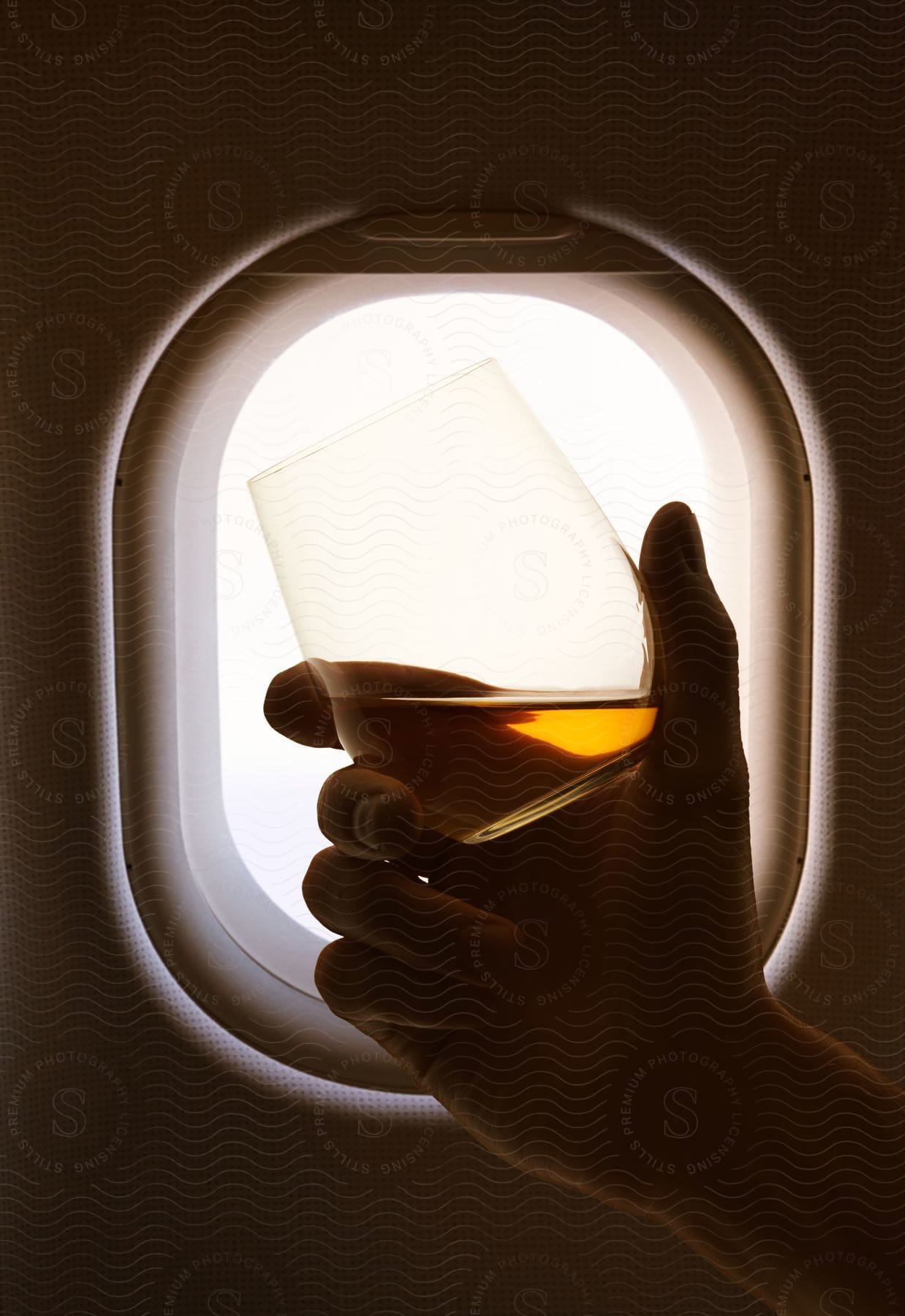 Hand holding alcoholic beverage against airplane window creating glowing effect