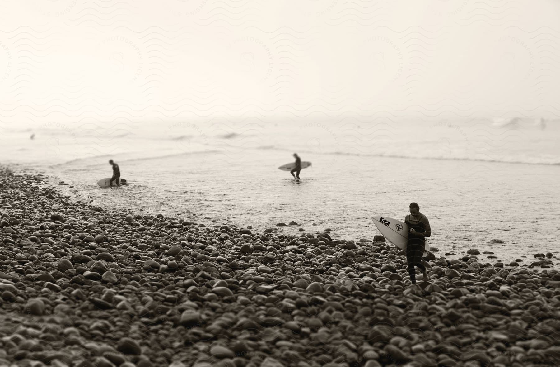 Three people walk on a rocky beach after surfing