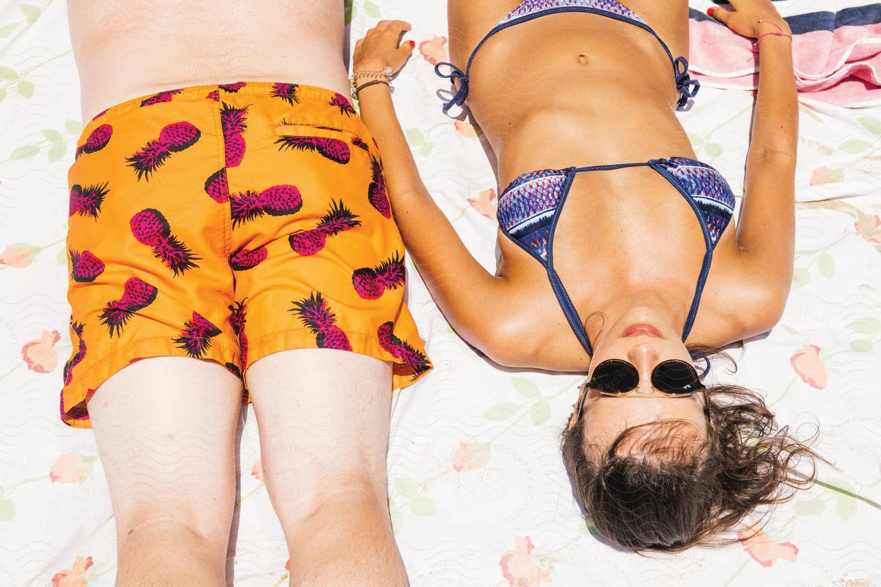 A Man And Woman Lay On A White Blanket In The Sun The Man Wearing Yellow Swim Trunks With A Pineapple Print And The Woman Wearing A Blue Bikini And Sunglasses