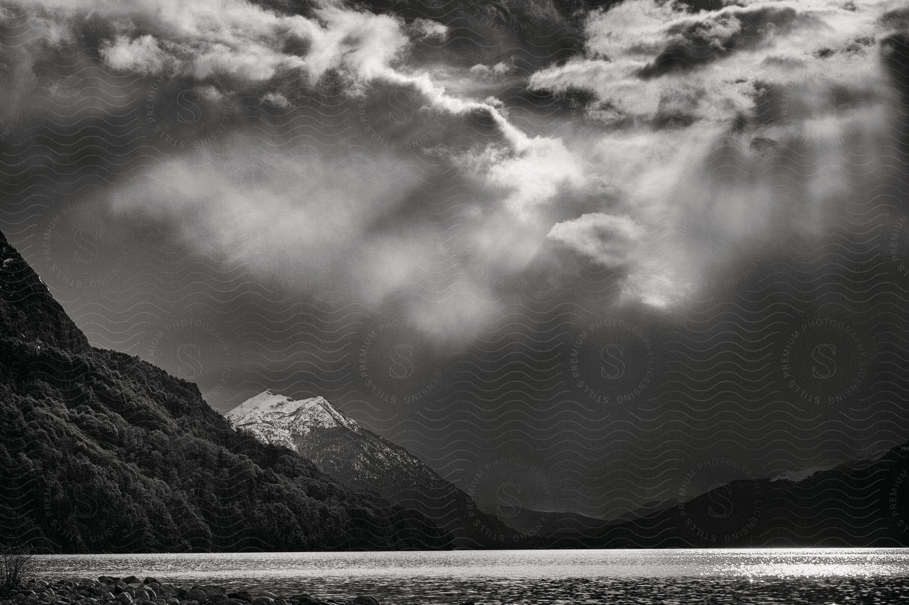 Stock photo of sunlight shines through clouds over a river at the foot of forested mountains and mountains in the distance