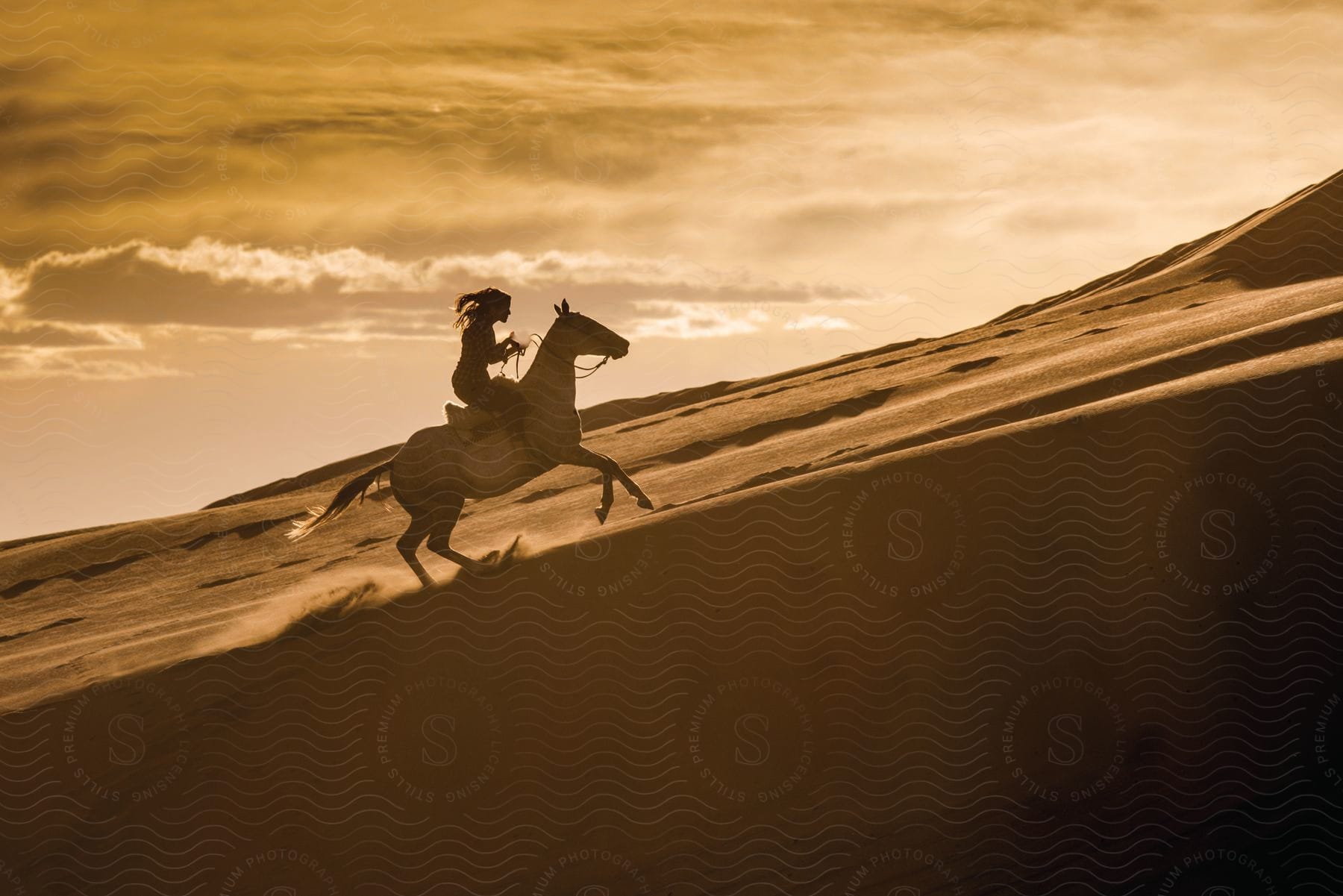 A woman riding a horse up a desert sand dune at dusk