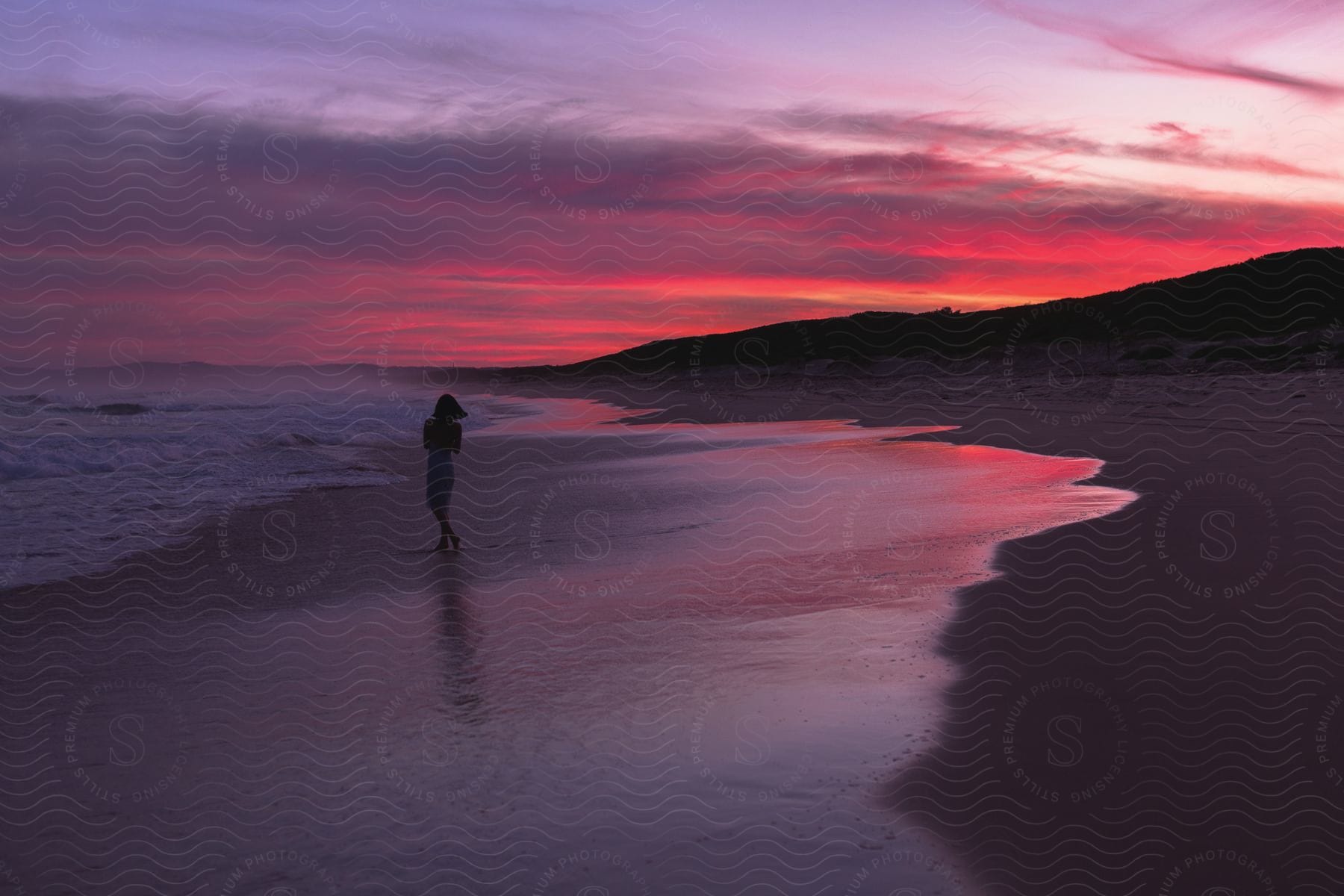 Woman walking on beach as waves roll into shore sun sets over mountains under colorful cloudy sky