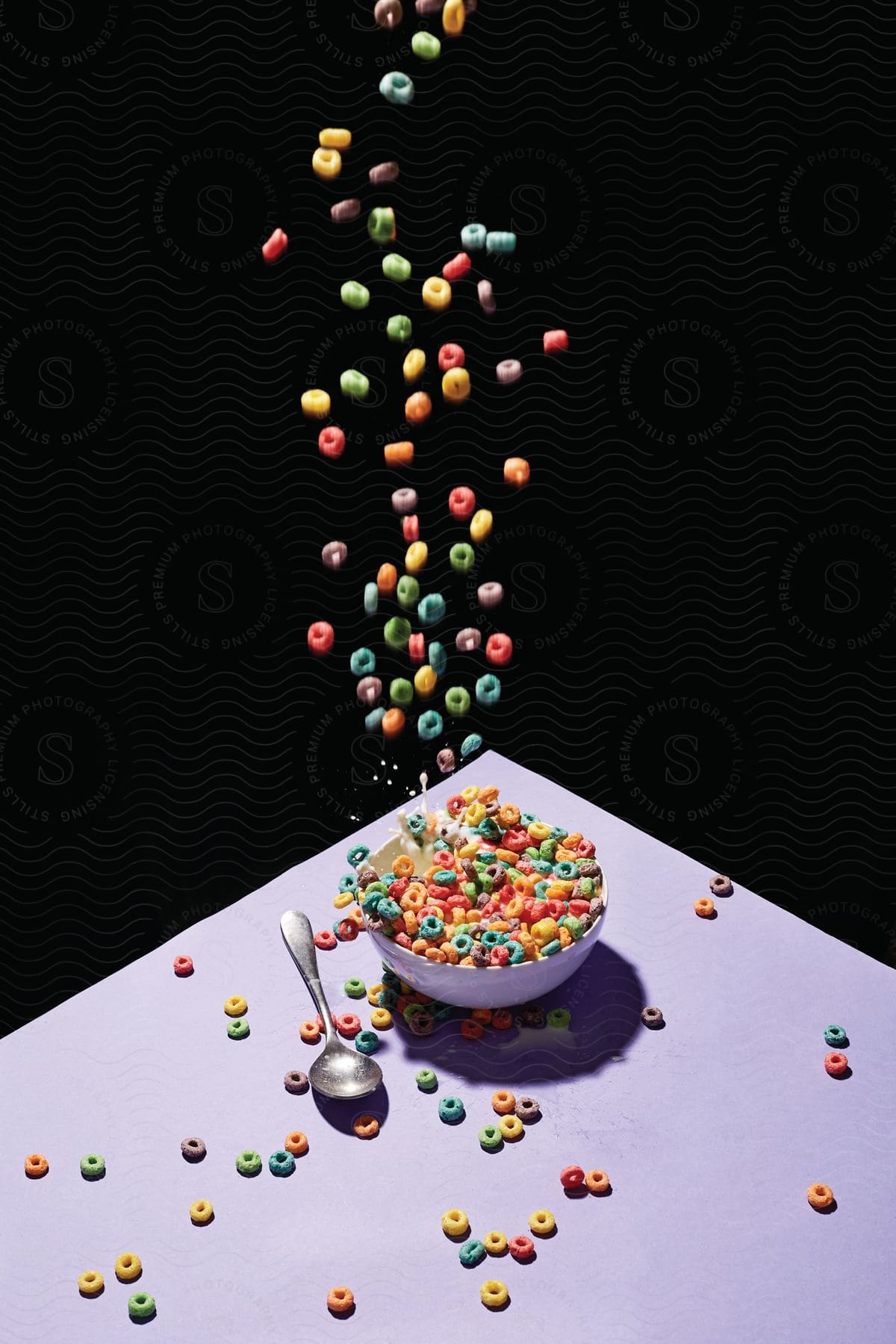 Fruit loops cereal falls into a bowl with milk from the air with some spilling on the table