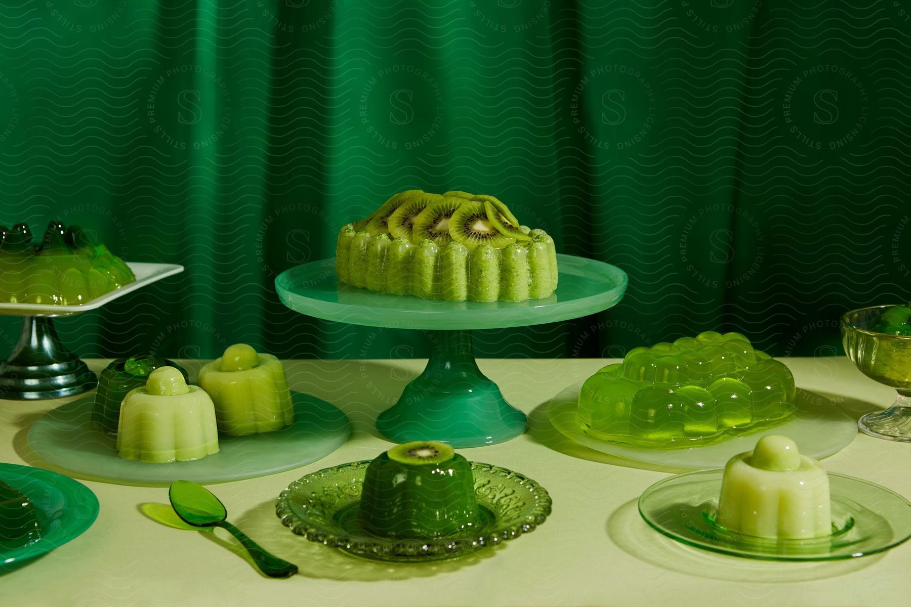 Green color scheme table presentation with kiwis and jello in a variety of green hues presented on different types of green glass platters on a white table in front of a green curtain