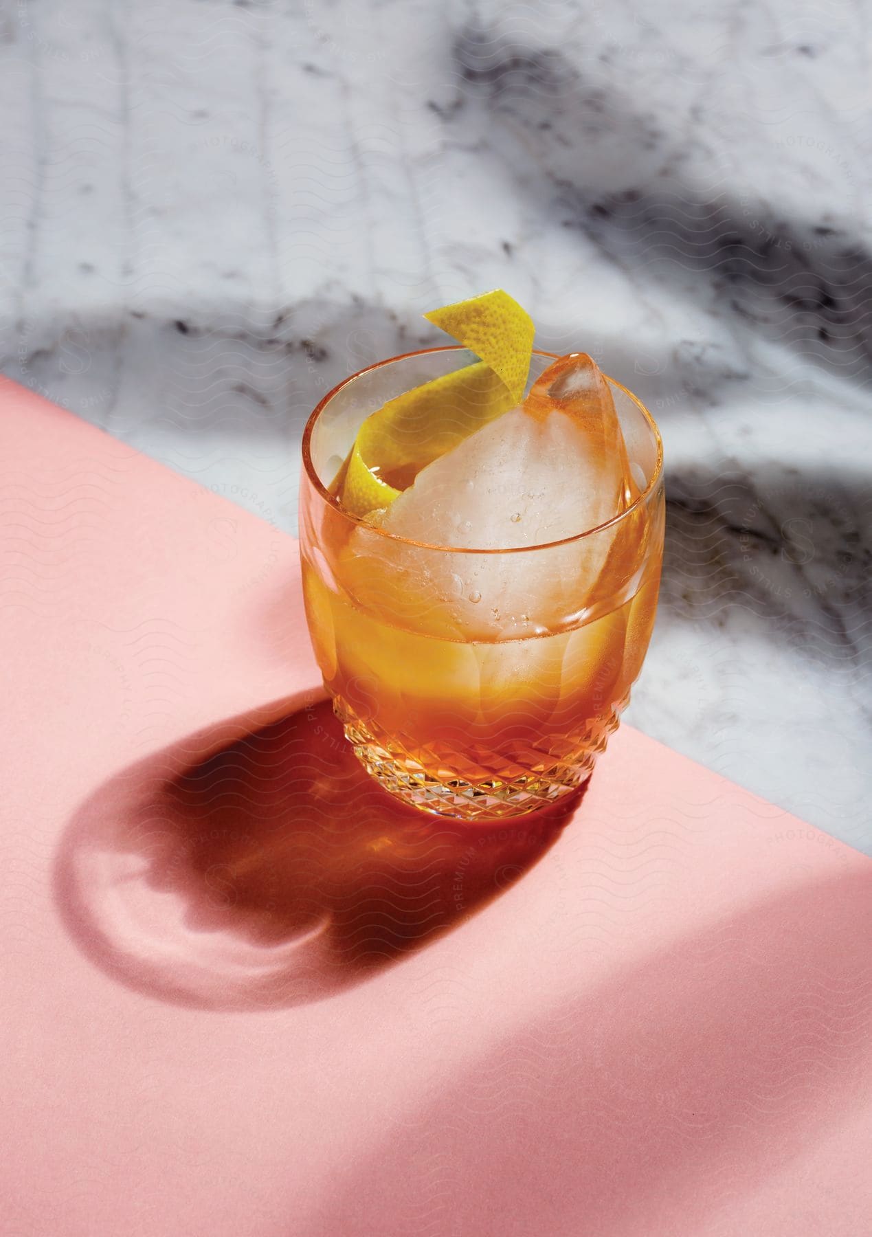 An alcoholic drink with a large ice cube and lemon peel garnish is placed on a pink table