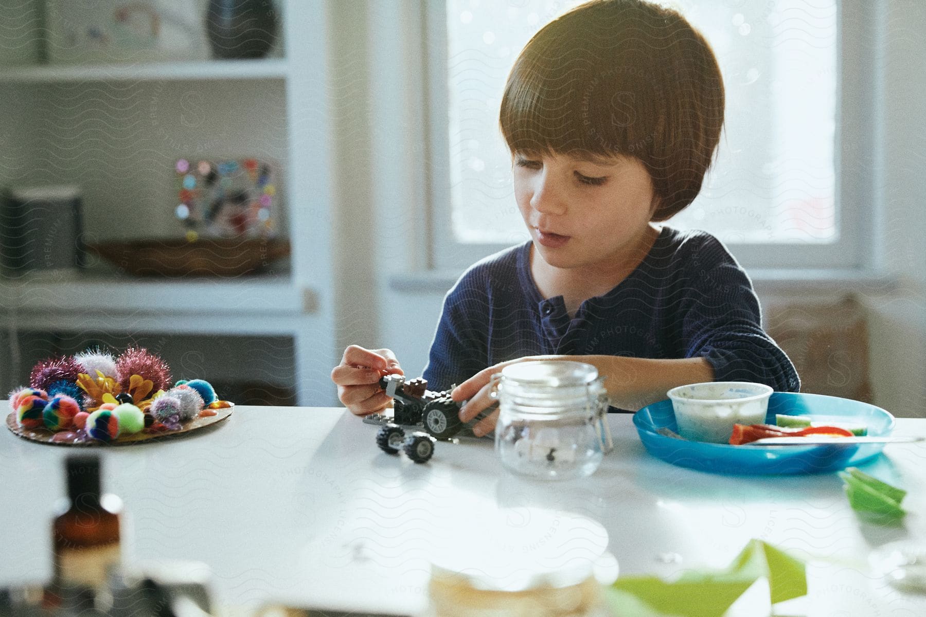 A boy plays with a toy car on a kitchen table surrounded by toys carrots celery and other kitchen items