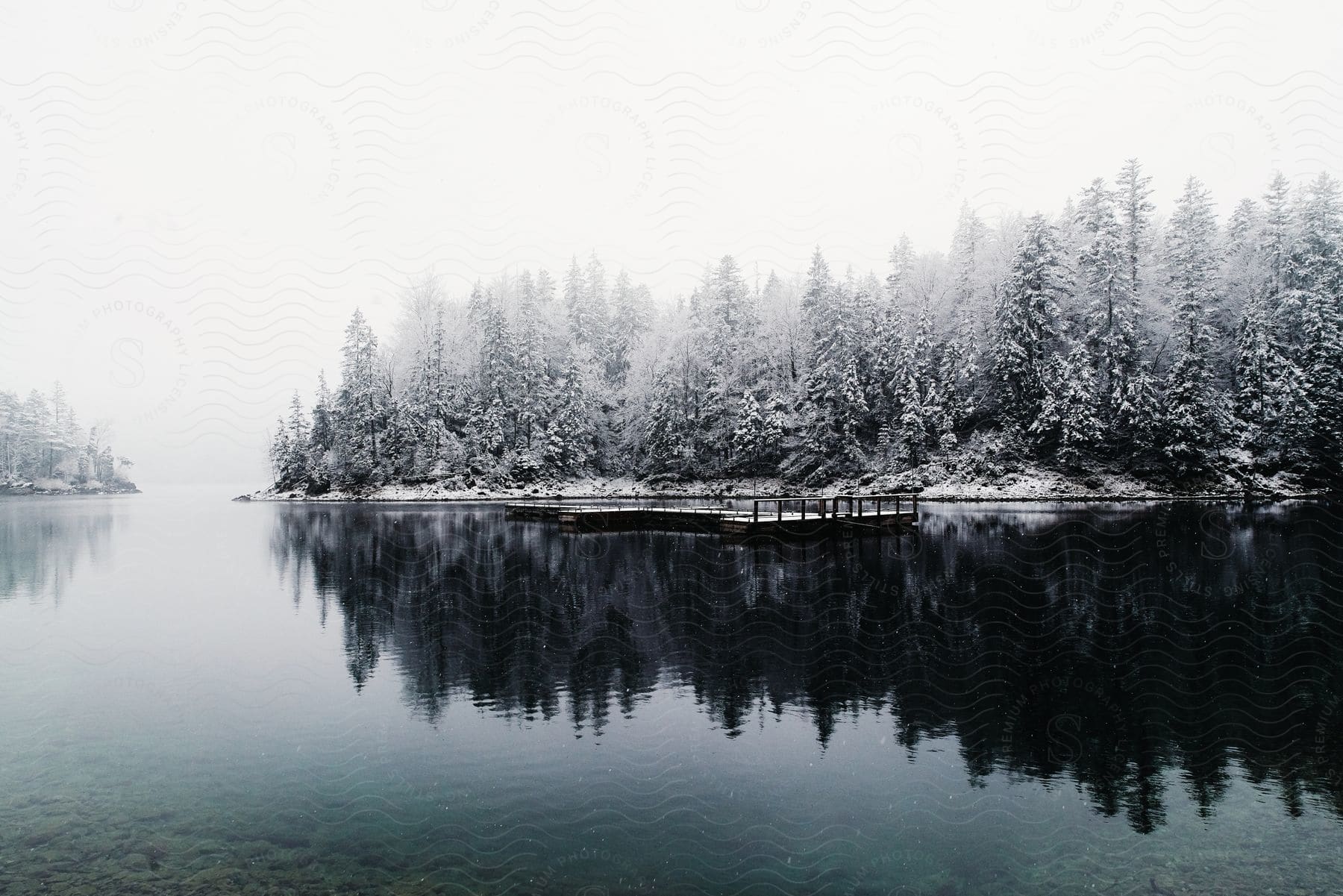 A snowcovered forest along the coast reflects on the water around a dock under a white winter sky