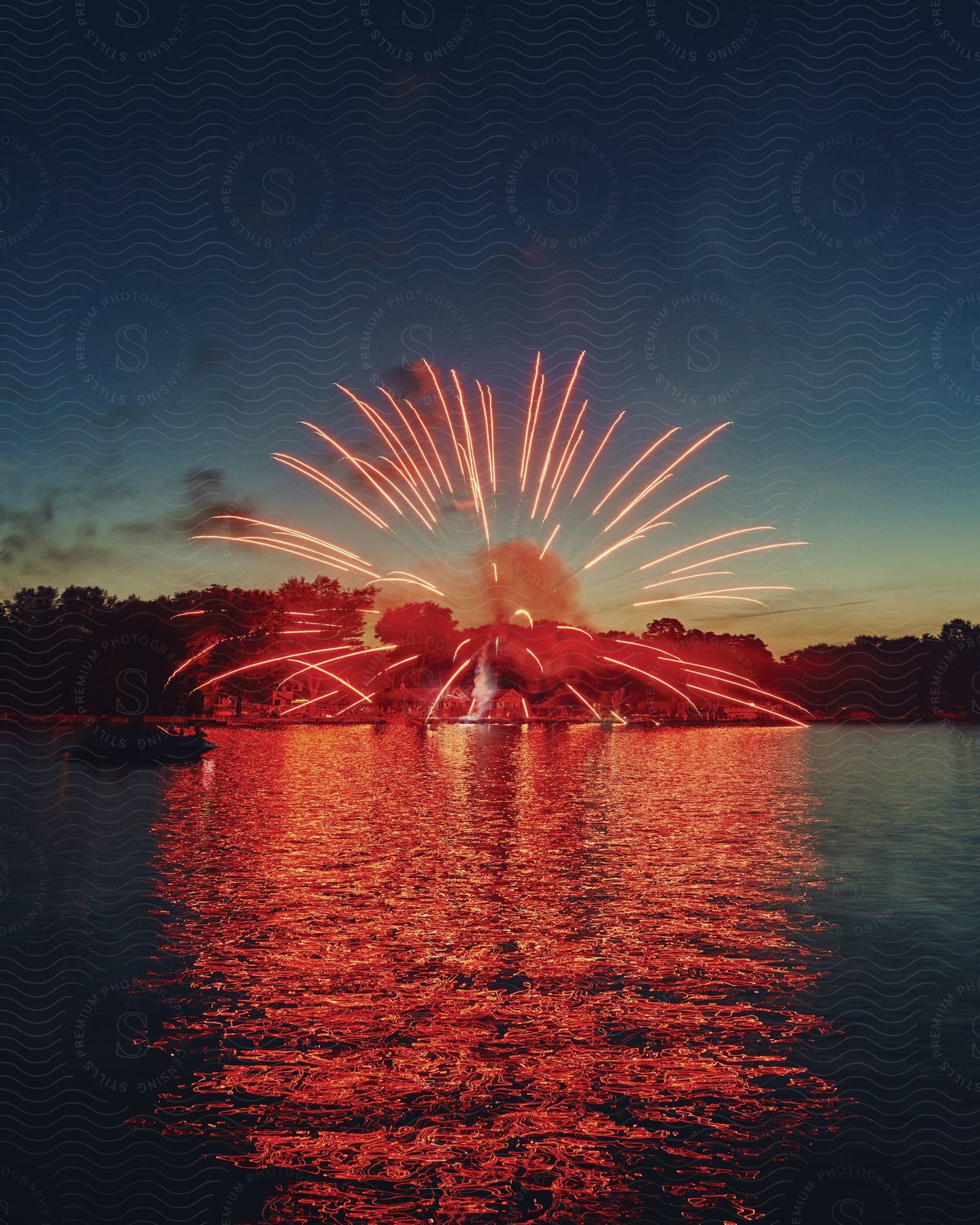 Vibrant bursts of red fireworks illuminate the night sky over a lake in a coastal town