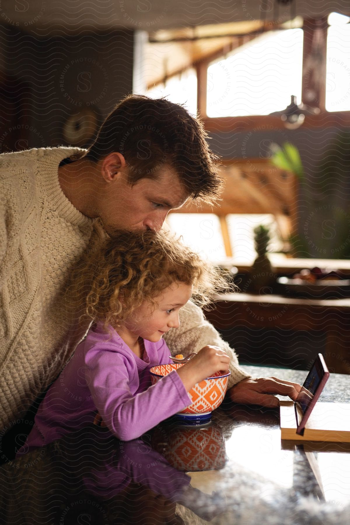 Stock photo of a father watches a tablet while his younger daughter watches while eating cereal