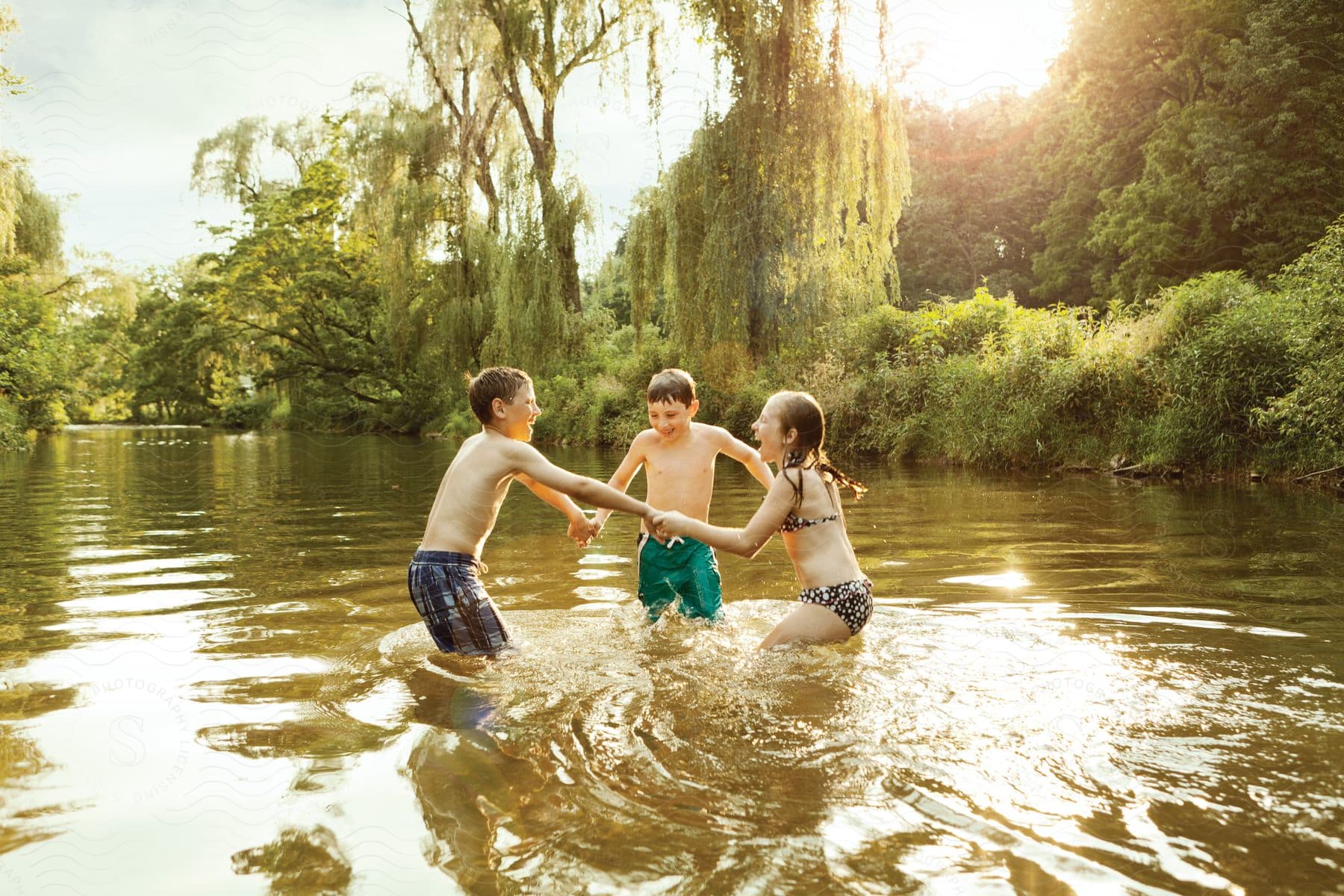 Three children holding hands in a circle jumping and laughing in a shallow river surrounded by trees on a sunny day