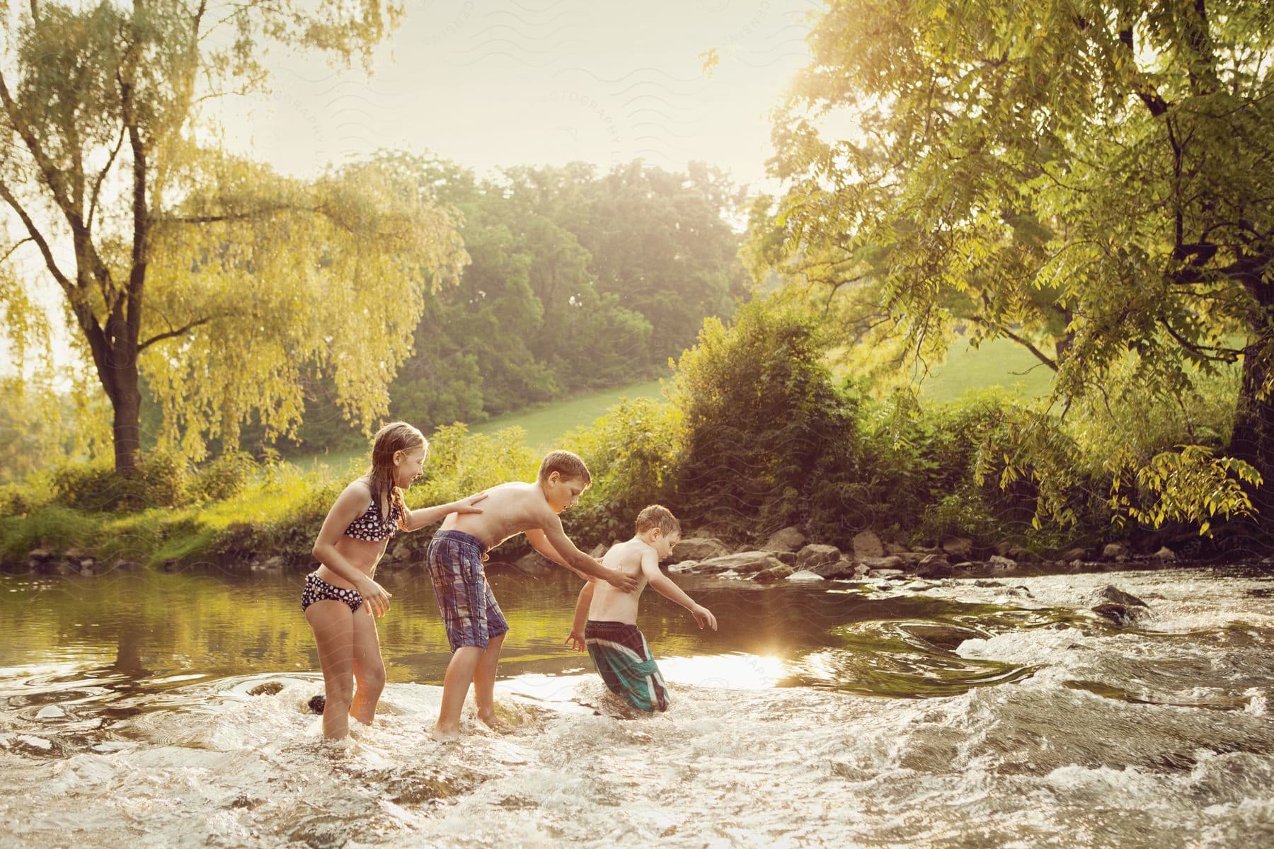 Three children walking through a river in swimwear holding on to each other