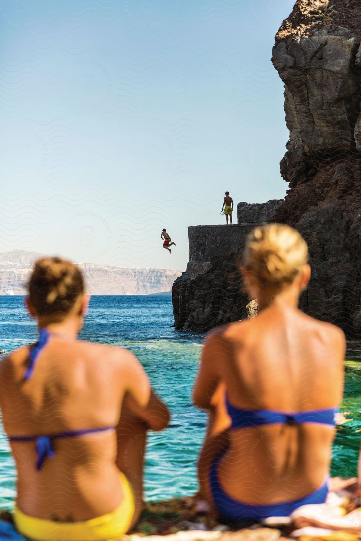 A person jumps off a cliff into the ocean with three friends watching