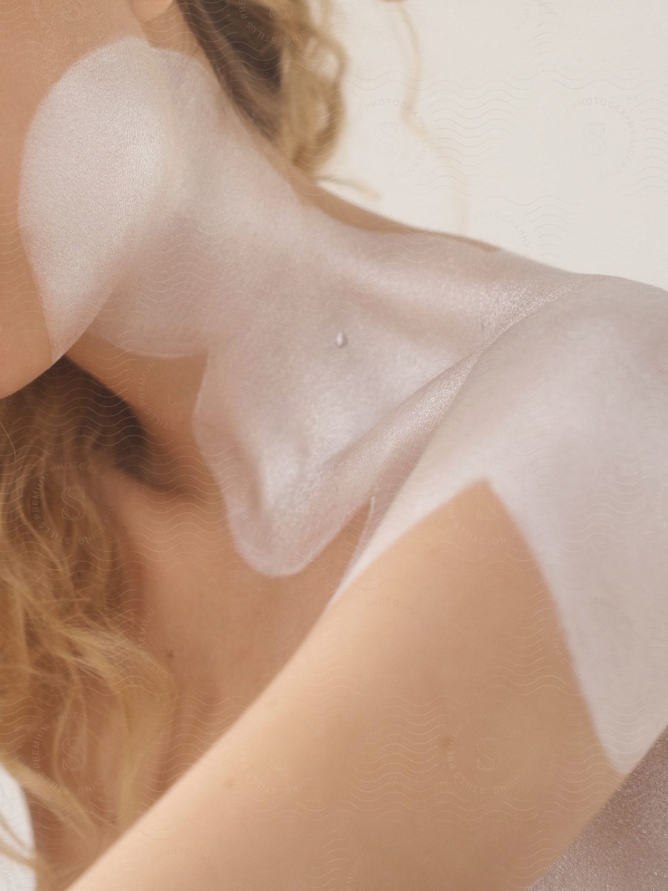 Blonde woman with gleaming paint on her shoulder and face poses against a studio background