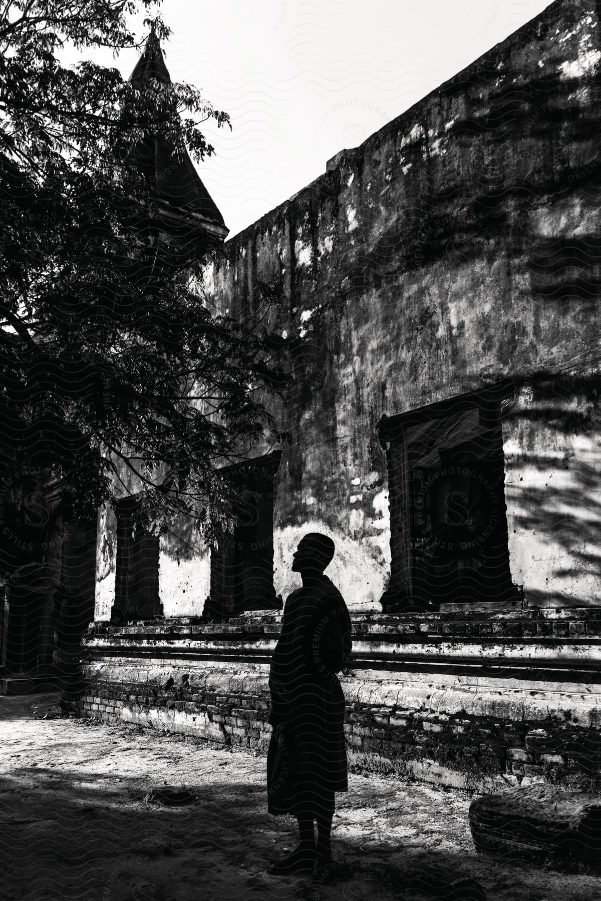 A person wearing a long garment stands in front of a tree next to a building