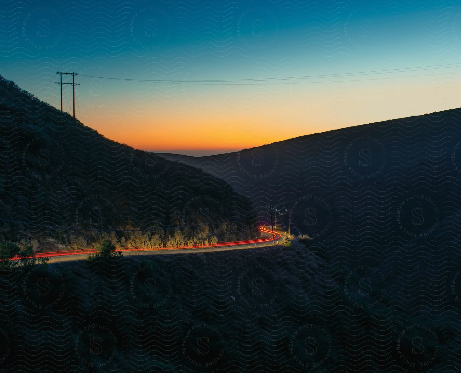 A road winds around a hill in a canyon illuminated by the setting sun power lines and passing vehicles