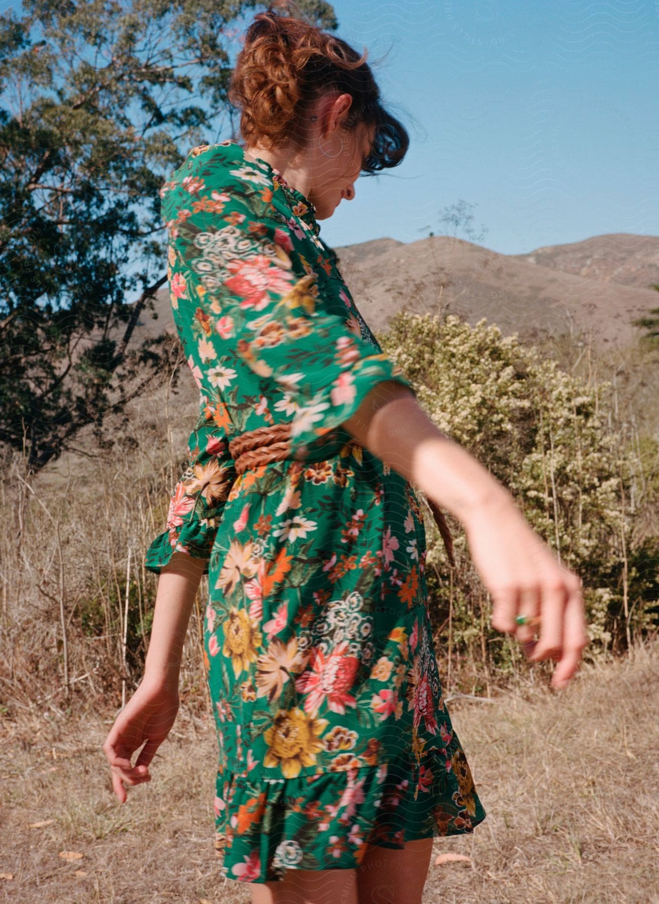 A woman wearing a green flowered dress stands at the base of dry mountains