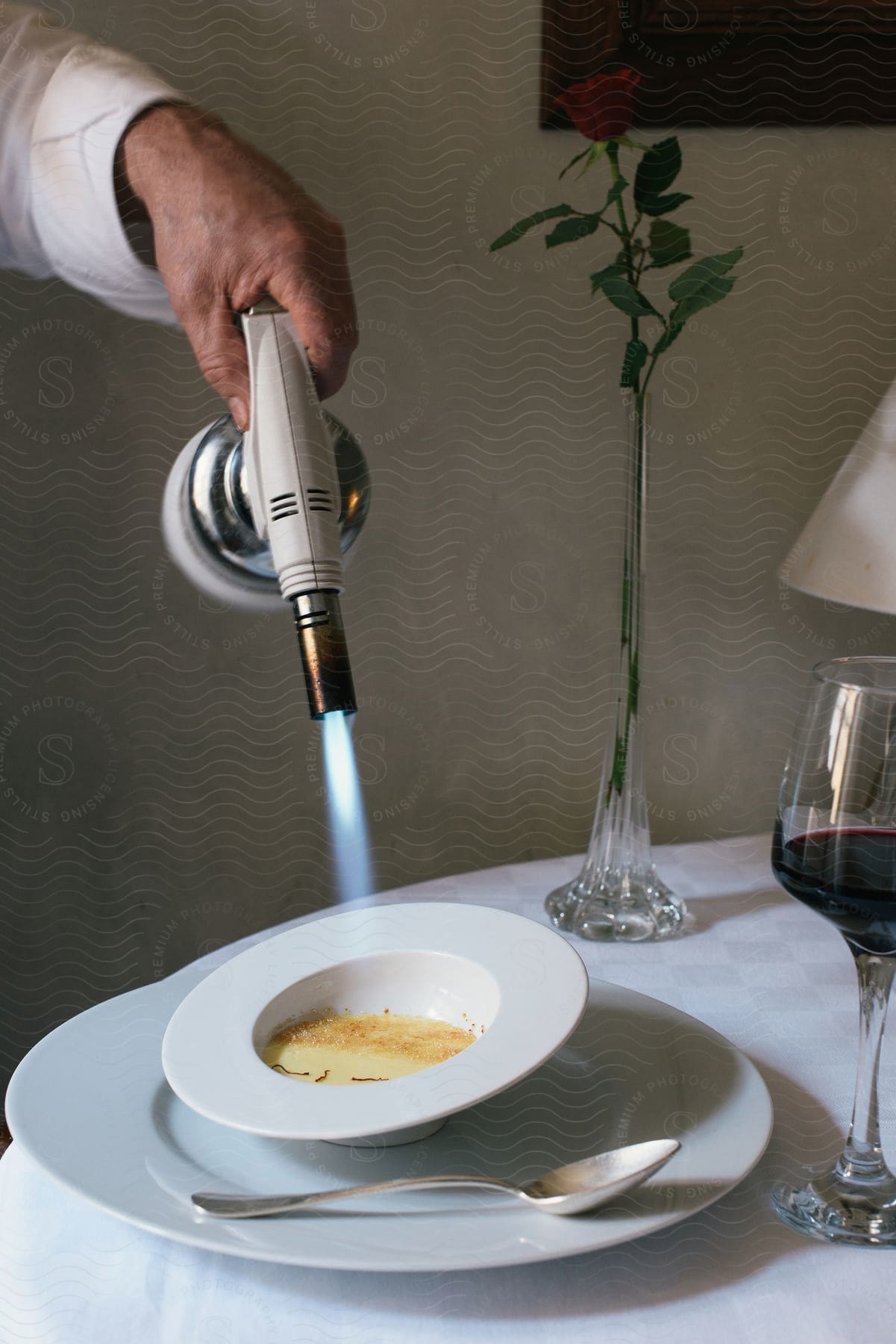 Stock photo of a waiter uses a blow torch to heat a custard in a bowl on a table with a glass of red wine and a rose