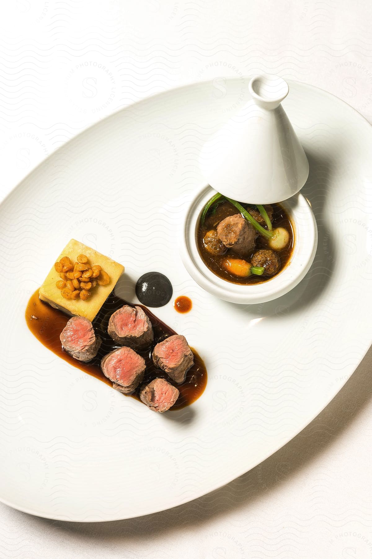 A white plate on a white table with a small bowl of brown stew a cup and meat rolls on top of a brown sauce