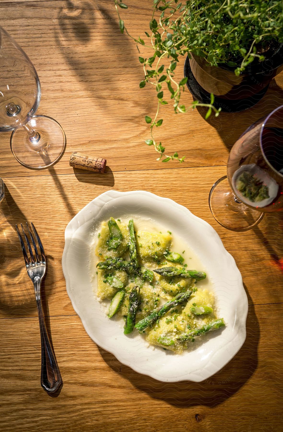 A dish of pasta and asparagus in a cream sauce surrounded by glasses a fork and a potted plant on a dining room table