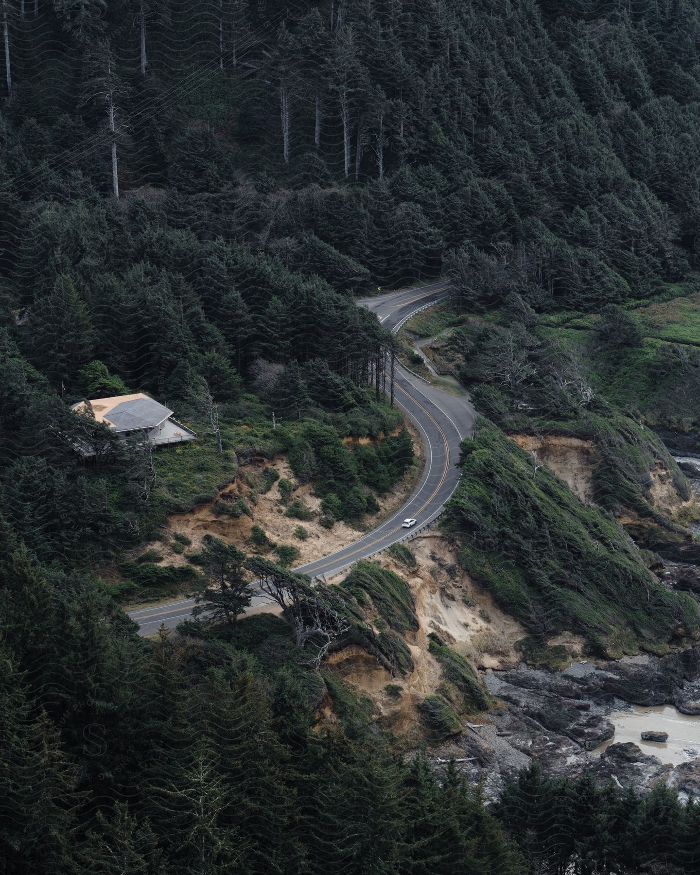 An automobile travels on a winding forest mountain road with a building at the foot of the mountain