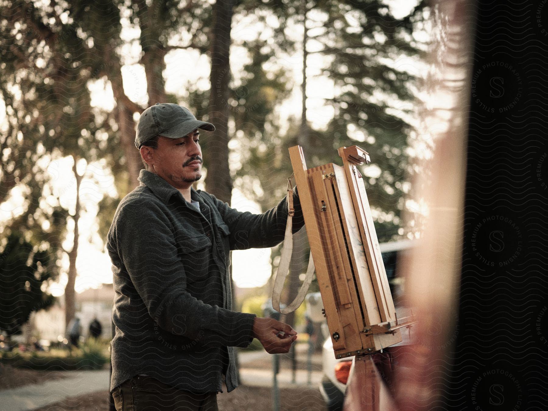 A latino man wearing a cap and long sleeve shirt is seen carrying an easel in a forest