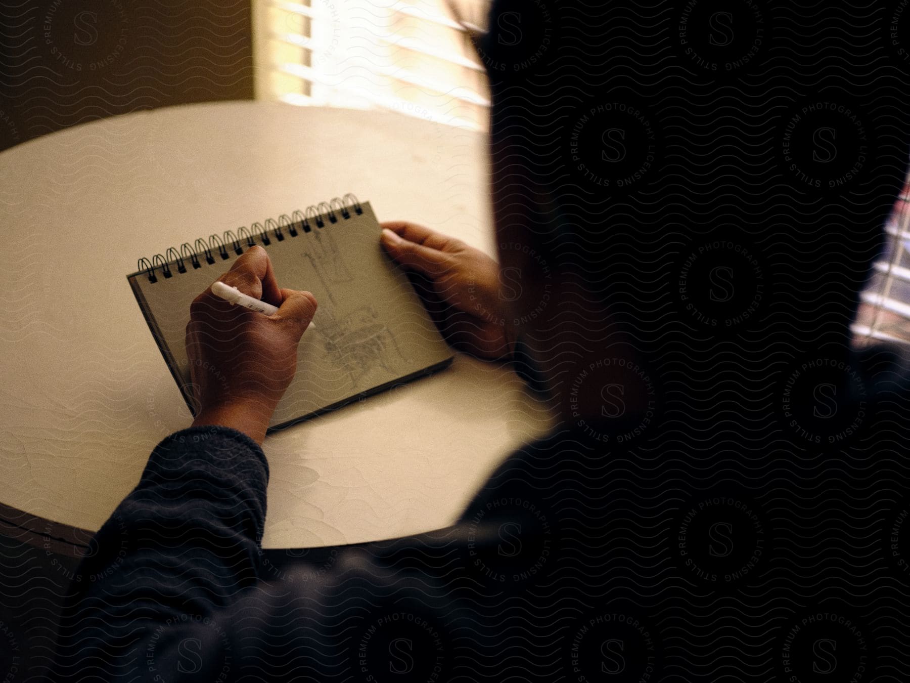 A man sitting at a table using a pen to draw on a notepad with light coming through the blinds