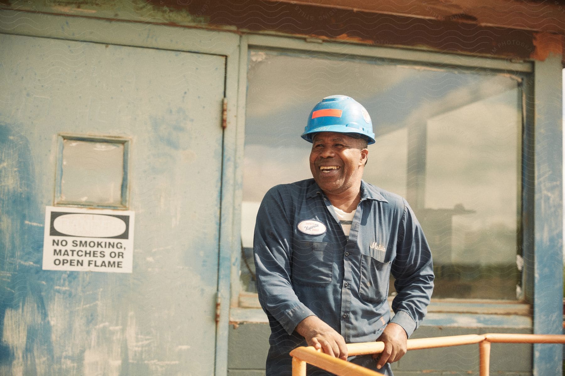 A worker in blue coveralls and hard hat standing outside holding a handrail and smiling in the sun