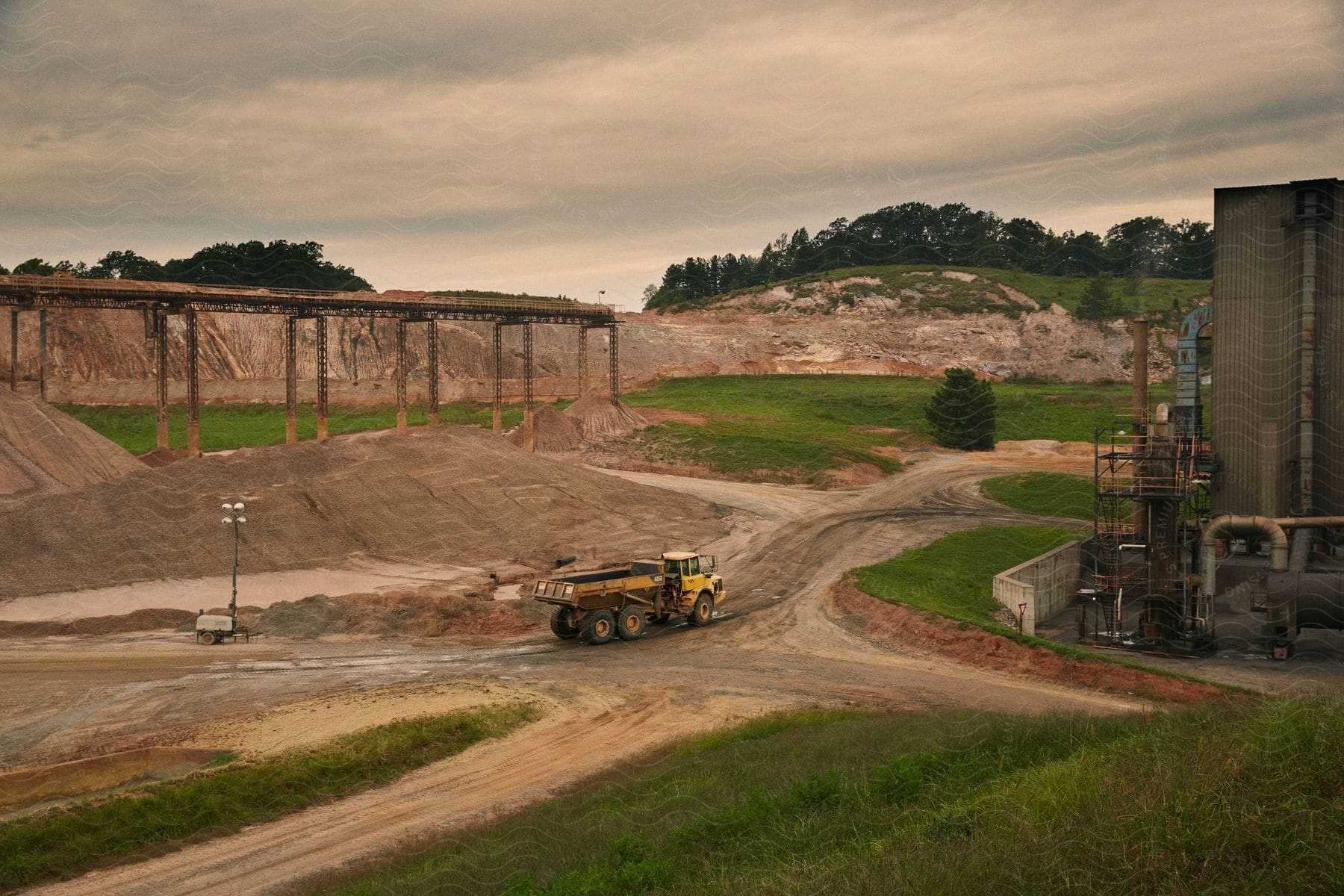 A truck drives on a dirt road near a pile of soil and machinery behind a building under a cloudy sky