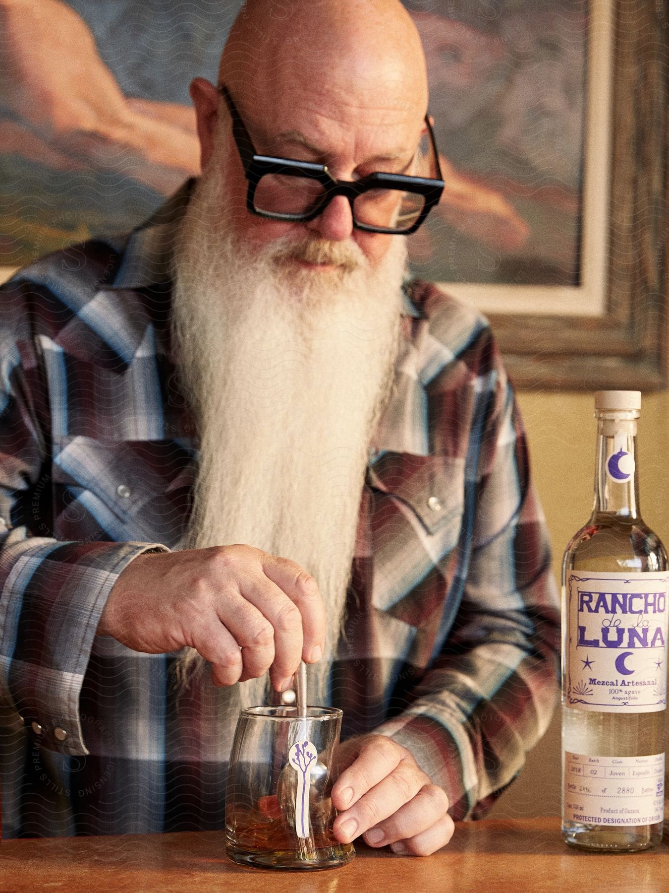 A bald man with glasses a flannel shirt and a long white beard prepares a drink on a glass with a bottle of mezcal next to him