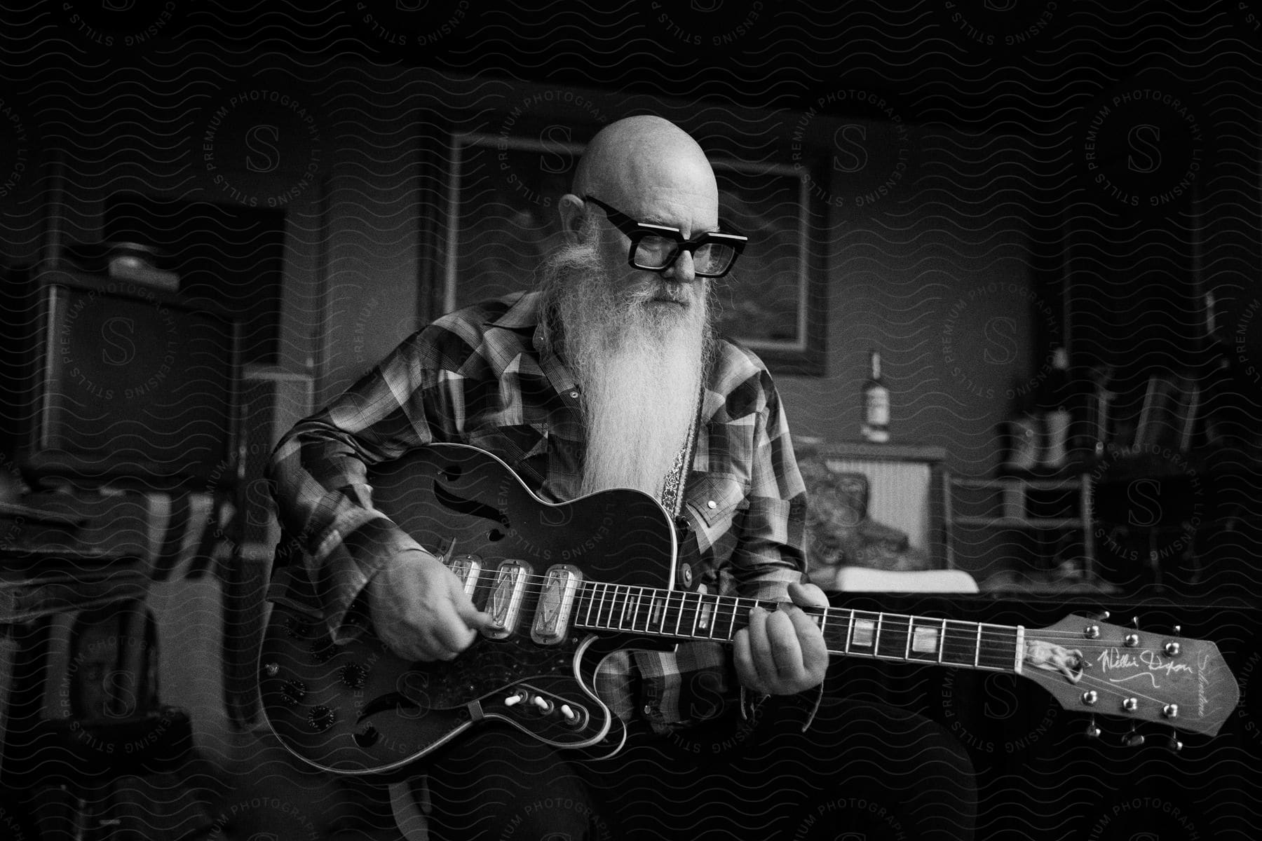 A man with a bald head white beard and sunglasses is playing the guitar in his home