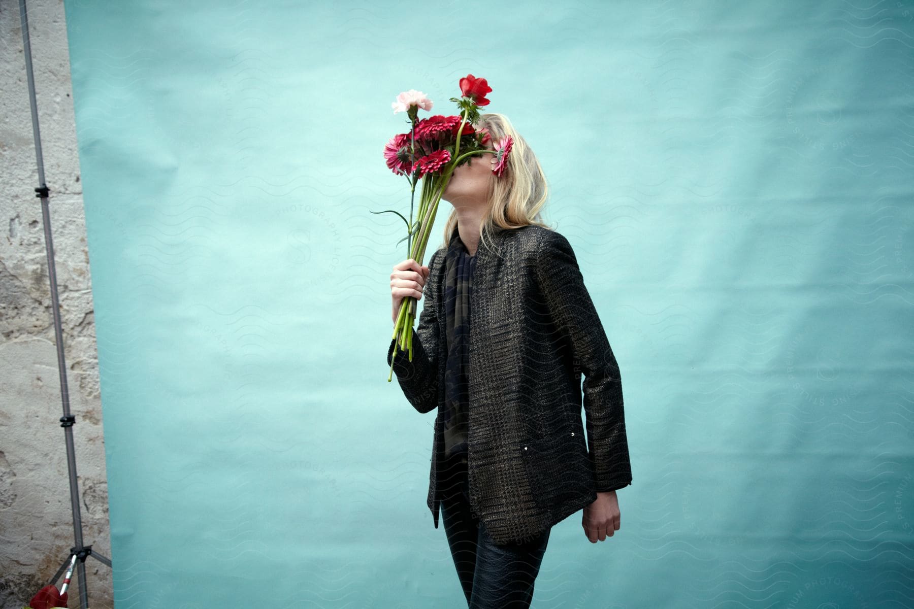 A blonde woman wearing a jacket and pants holds a bouquet of flowers in front of her face