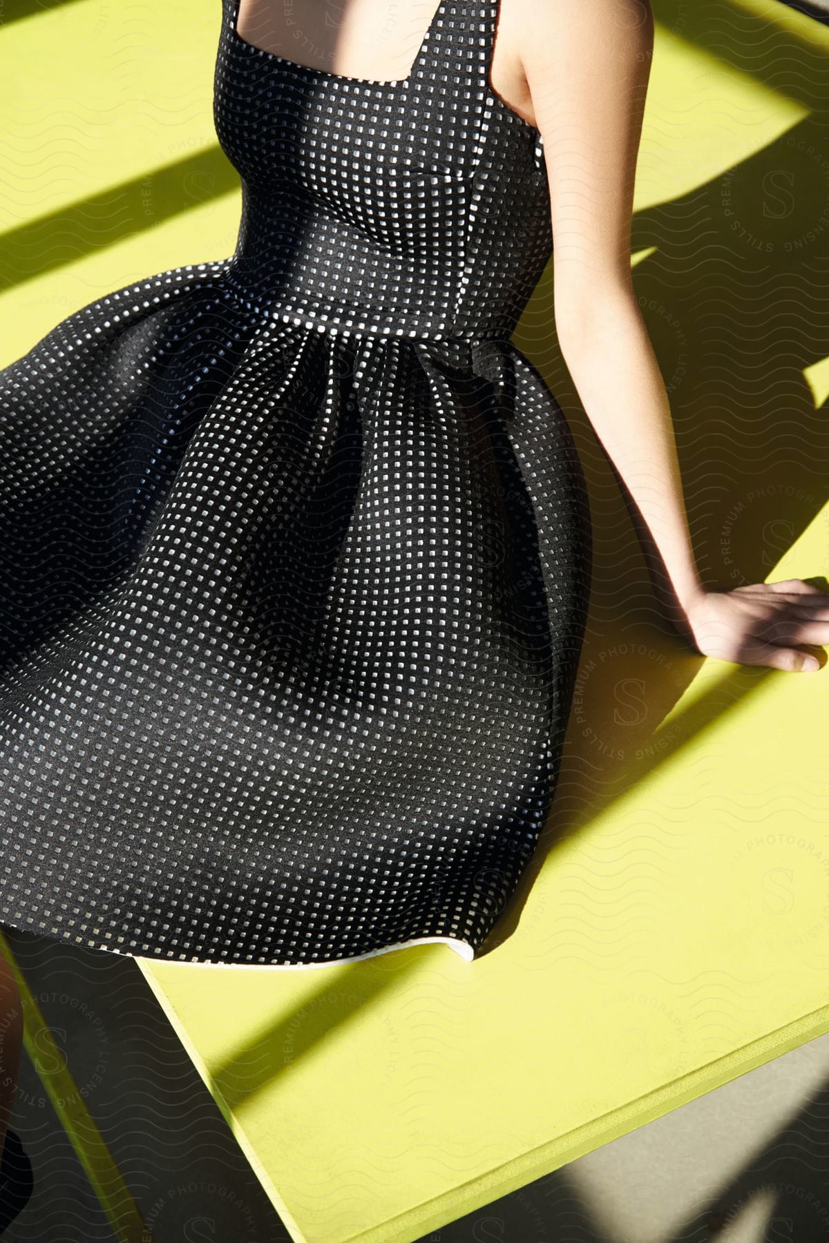 A young woman leans against a yellow table top while wearing a black and white polka dot dress