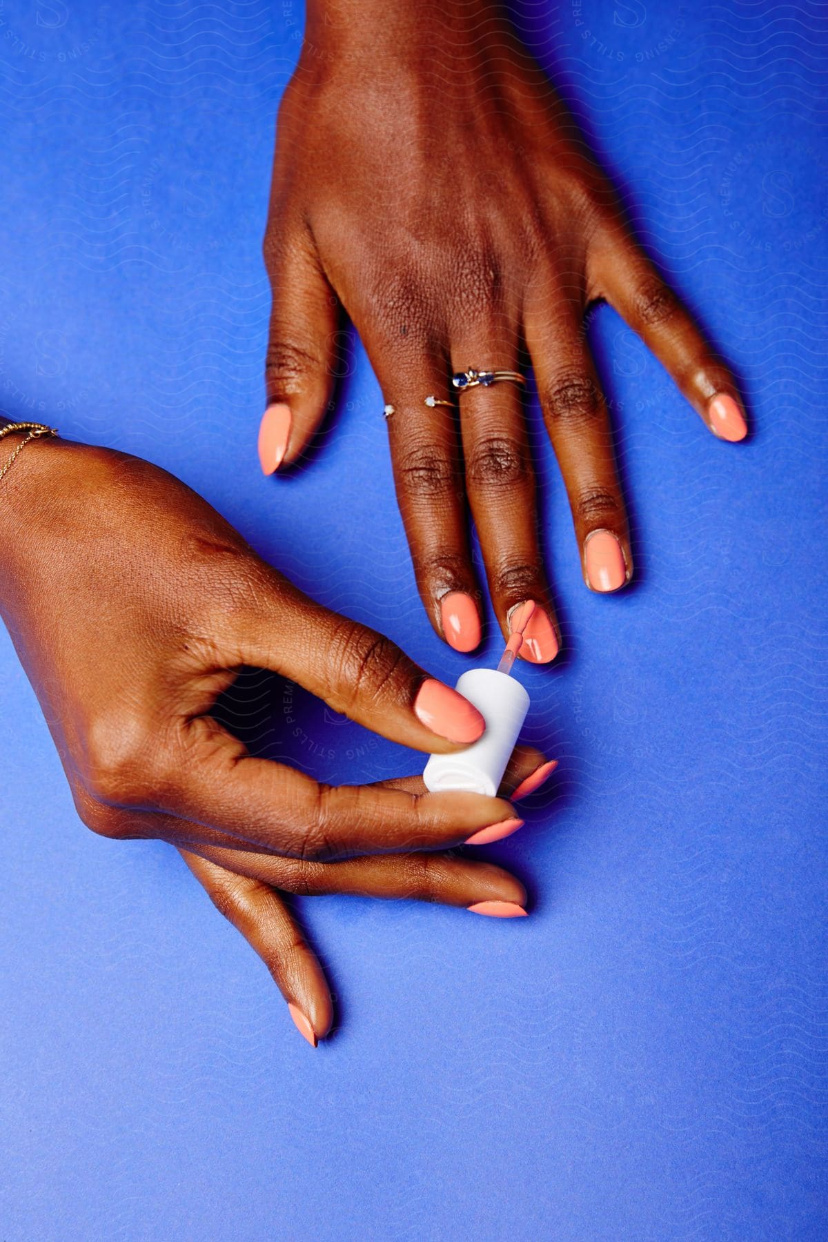 A woman applies pink nail polish to her nails with a brush while wearing a ring on her finger on a blue surface