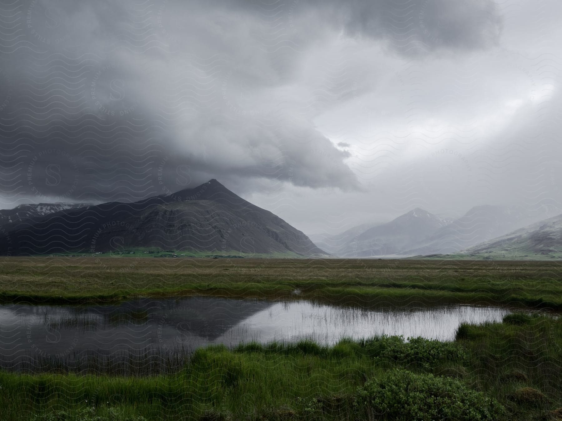 A dark cloud hovers over a mountain while a lush green grassland stretches out in the foreground with a small lake adding to the scenery