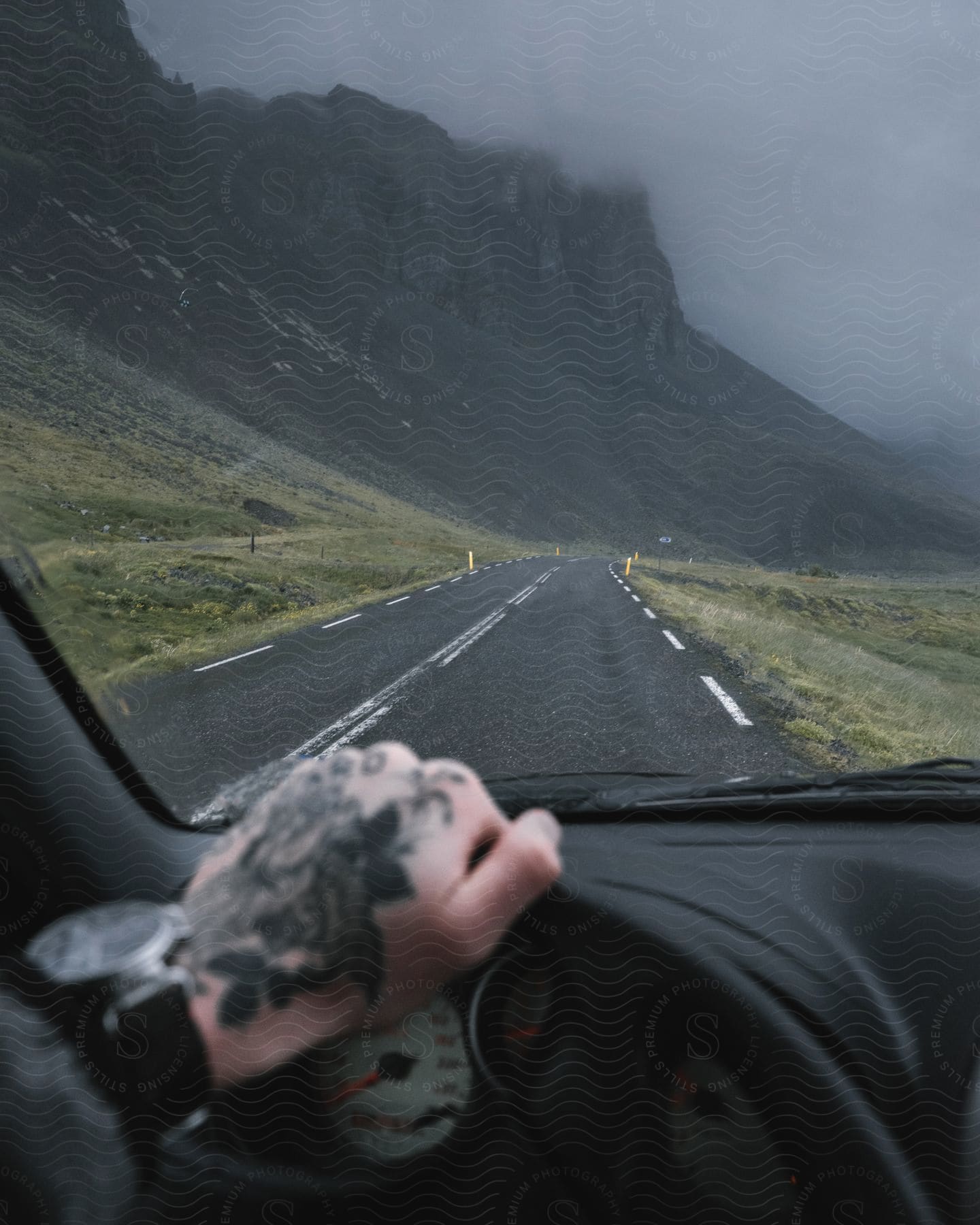 A drivers hand and watch are visible in the frame as they view mountains and a road from the drivers seat of a car