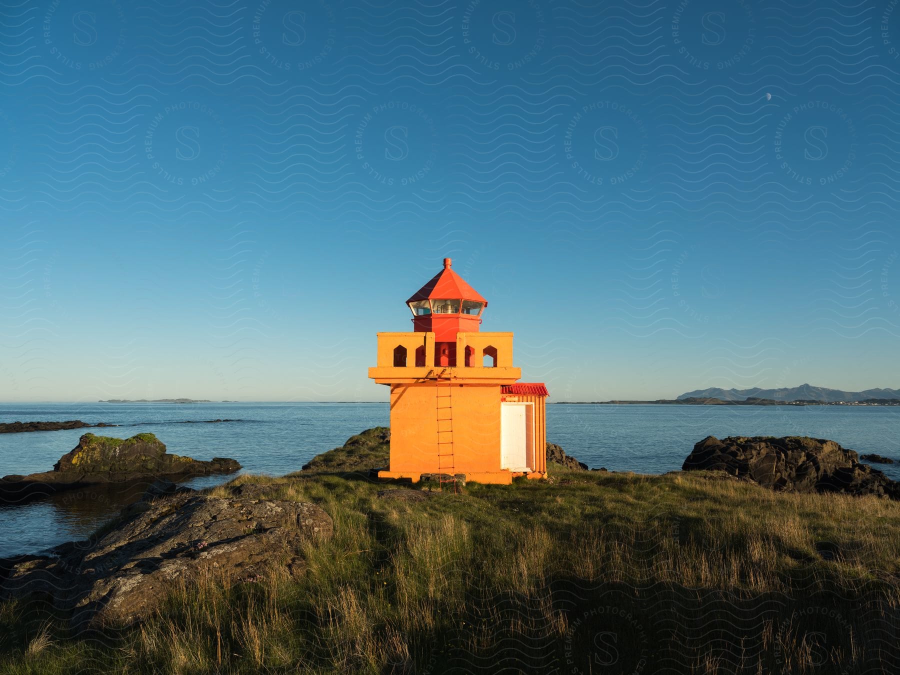 An orange and red lighthouse stands at a rocky grassy edge next to a bay at dusk