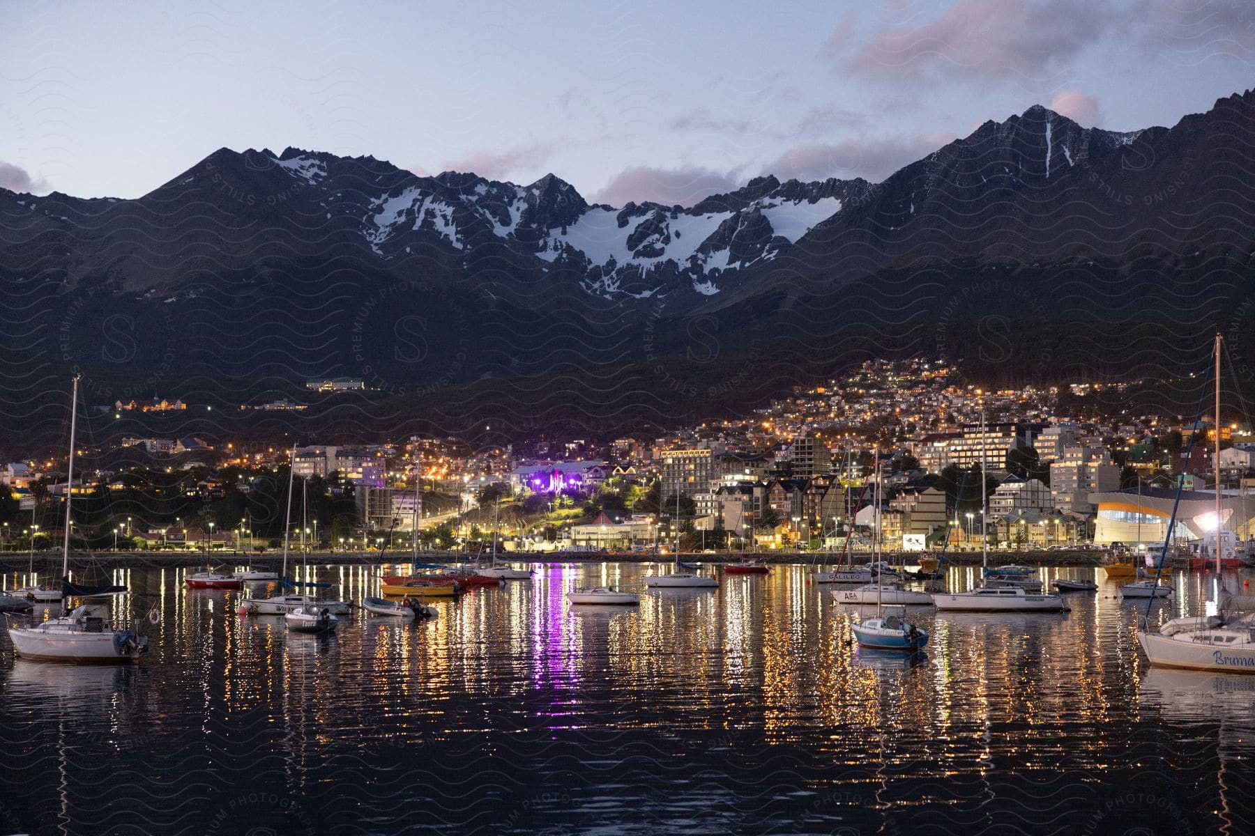 A city with boats anchored near shore and a snowy mountain in the distance seen at night