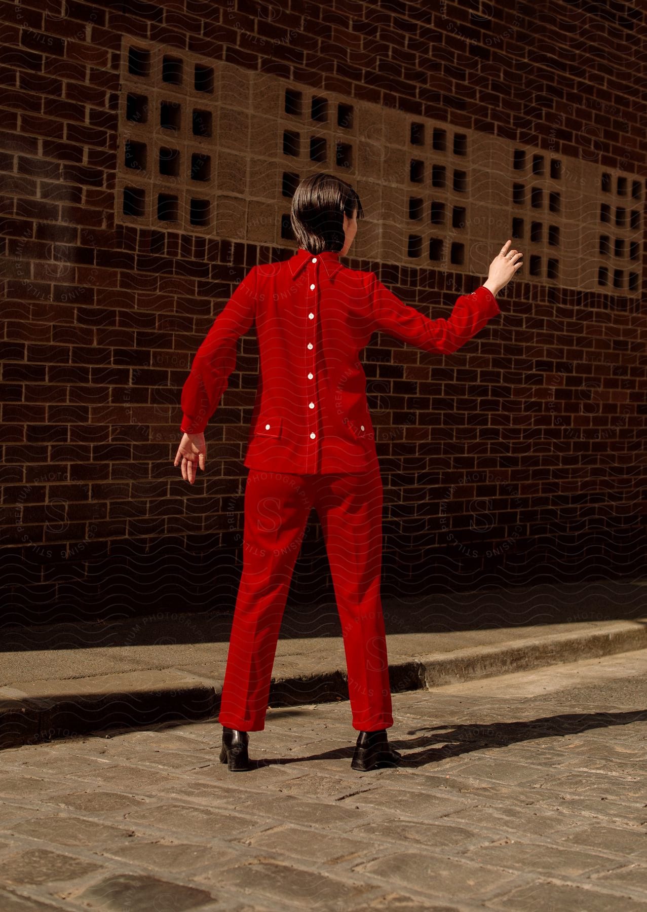 A person wearing a suit and coat stands against a brick wall making a gesture with their sleeve