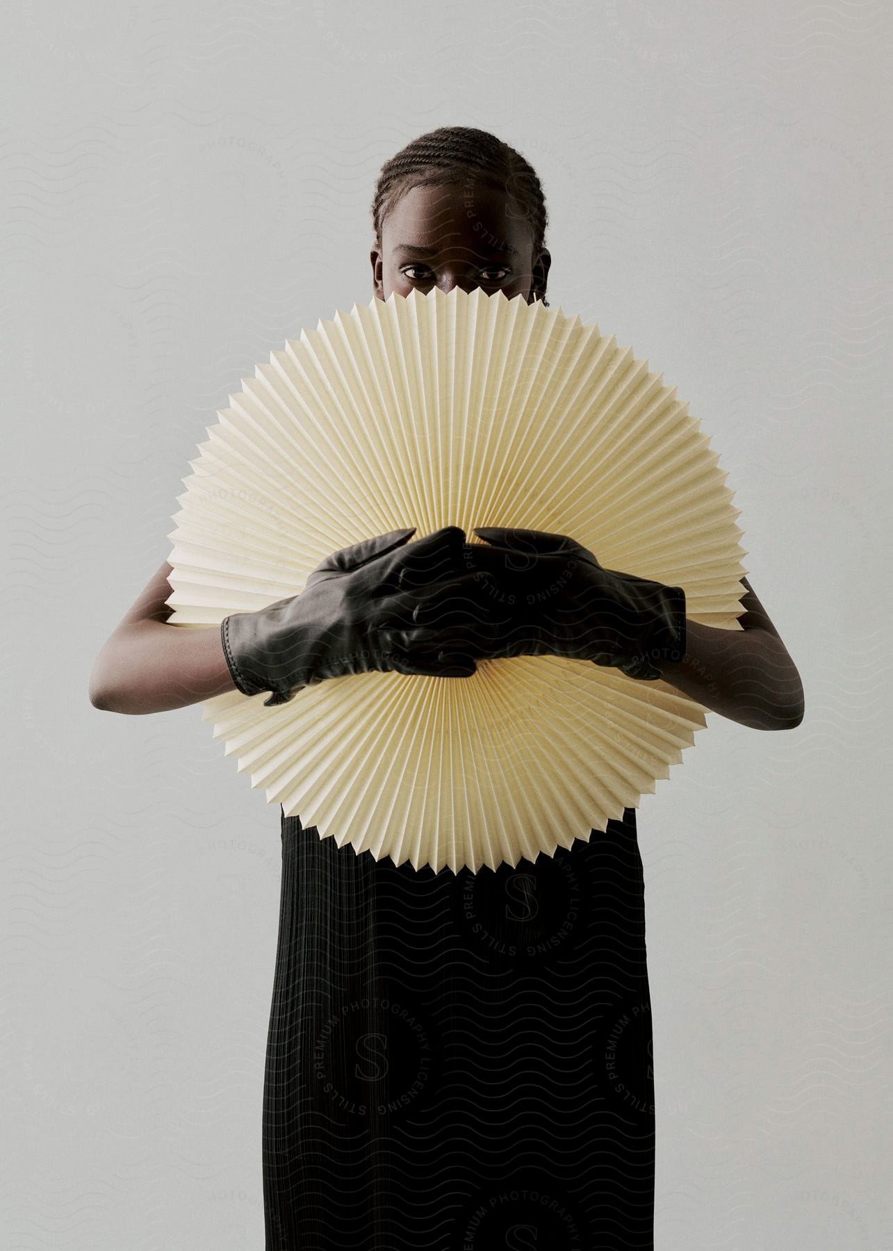 A black woman wearing a black dress and black gloves hides half of her face behind an object