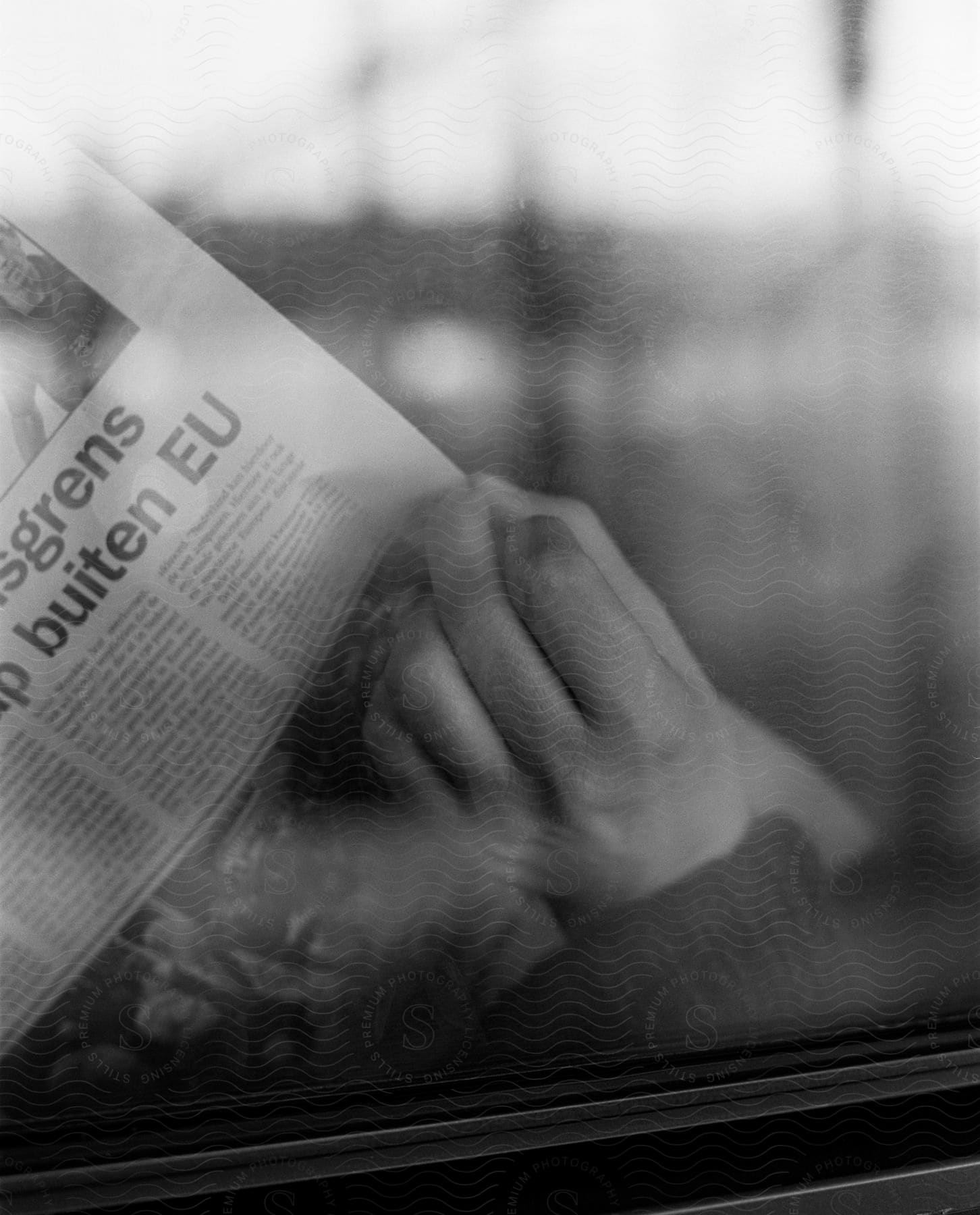 A blurry reflection on a window and a close up of a persons hand holding a newspaper on the other side of the window